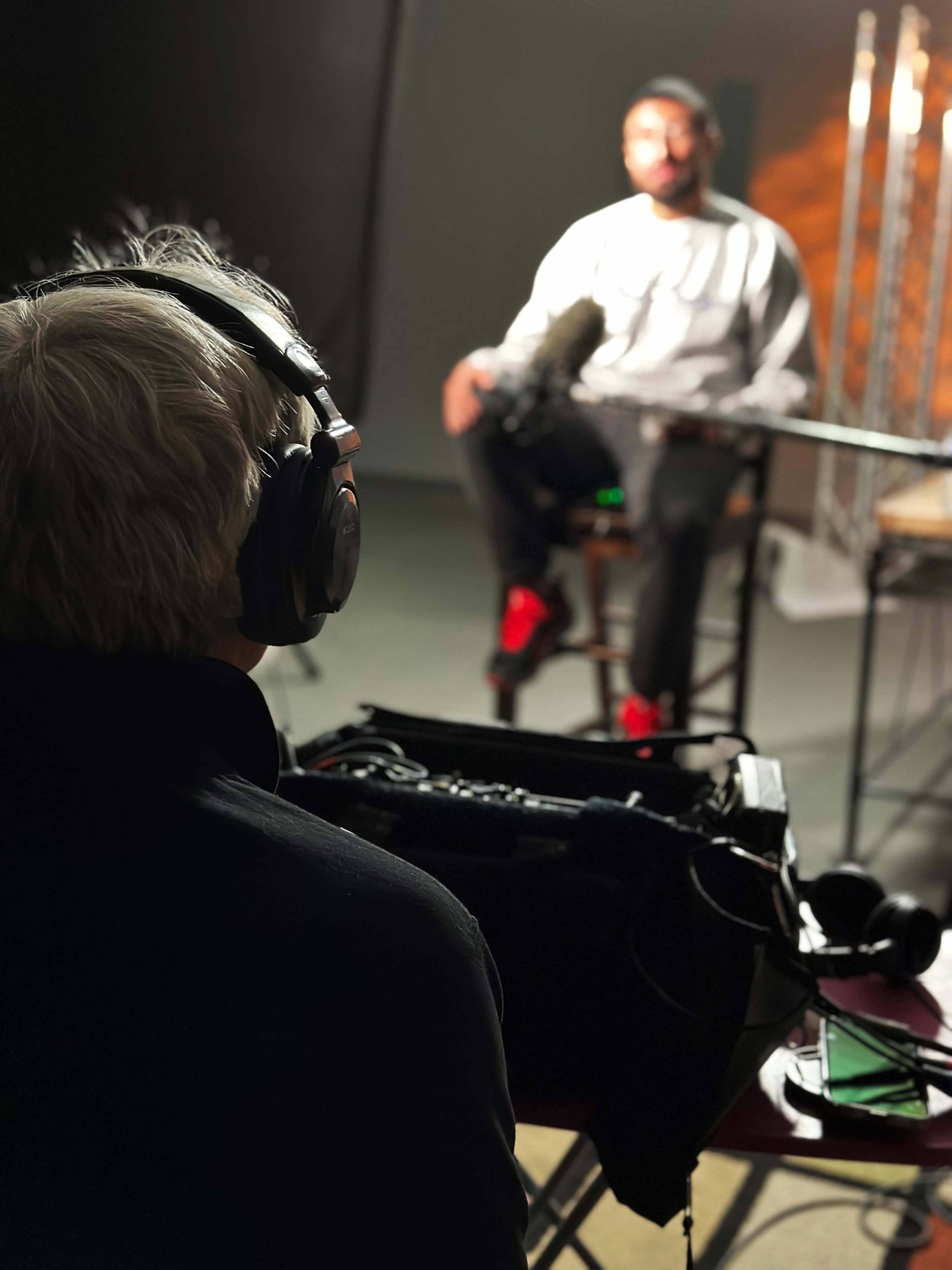 A person wearing headphones operates audio equipment while another person sits on a stool in front of a microphone.