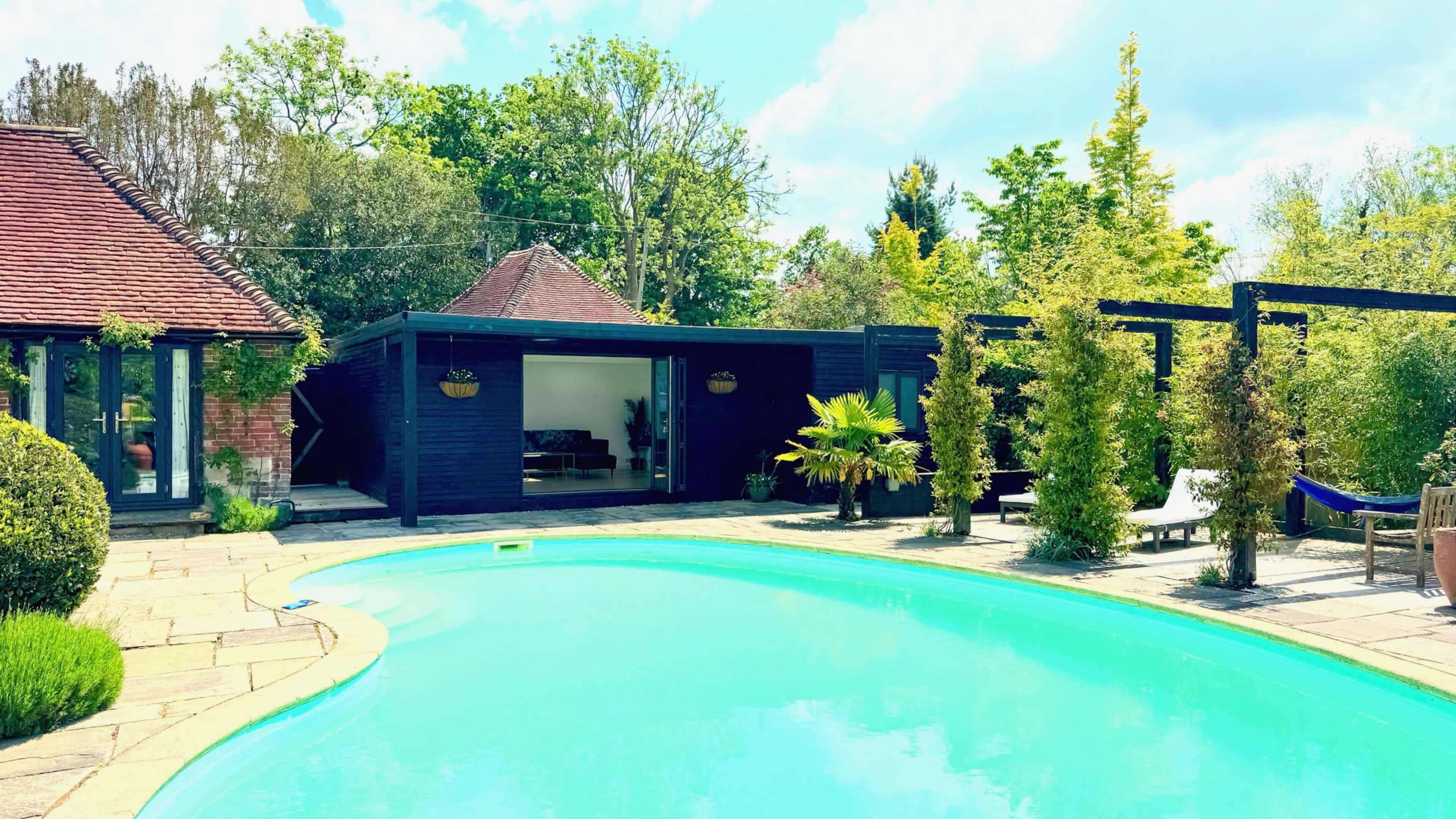 A rectangular swimming pool is surrounded by neat stone patio and lush greenery, with a dark wooden structure and landscaped area adjacent to it.