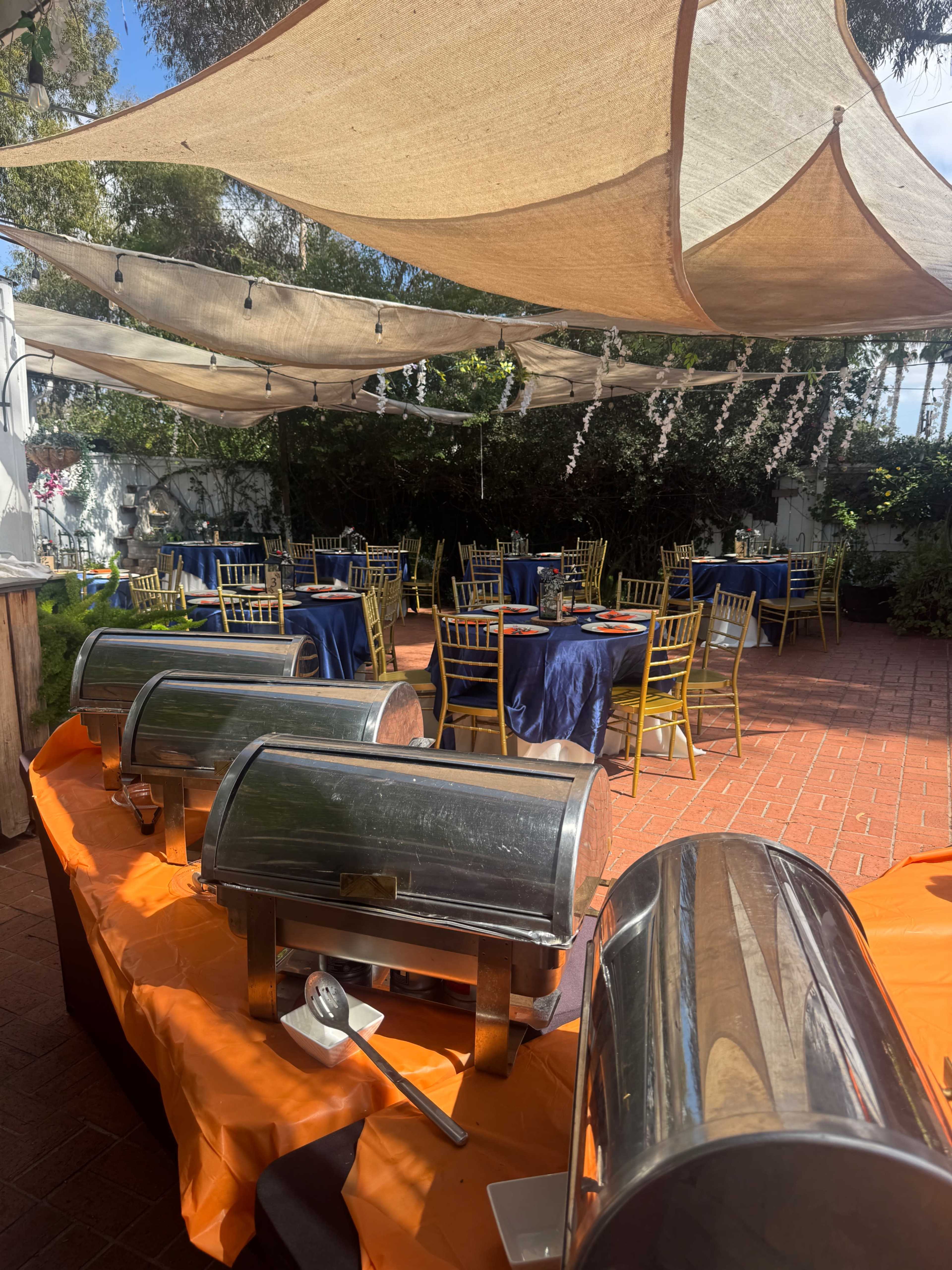 The scene shows a sunny outdoor event space with several buffet warming trays in the foreground and elegantly set tables with navy blue tablecloths and gold chairs in the background.
