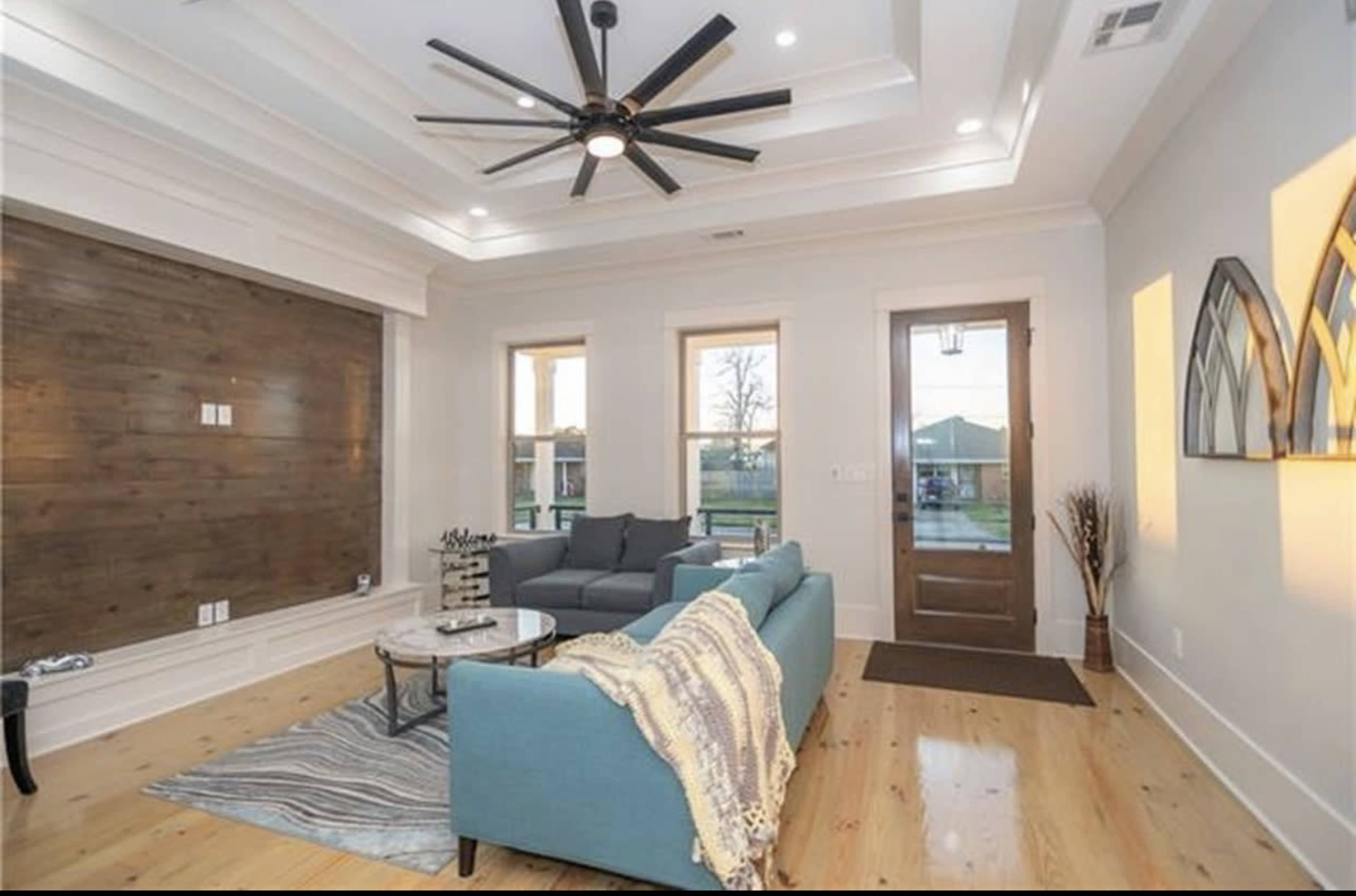 The image shows a modern living room with a blue sofa, a coffee table, and a large ceiling fan, complemented by natural light from the windows and a wooden accent wall.