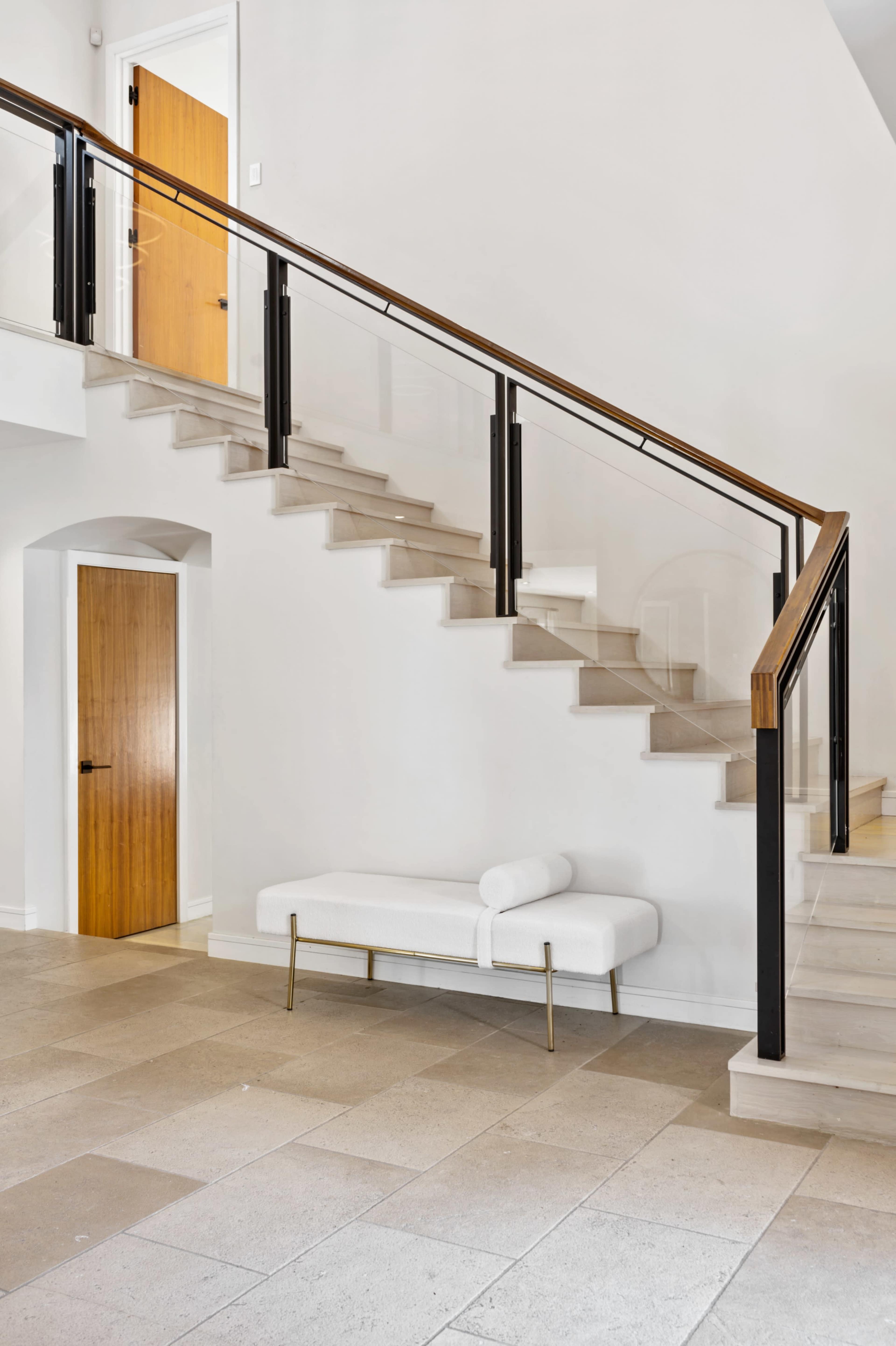 A modern interior with a wooden staircase featuring glass railings, a white bench, and a door on the left side.