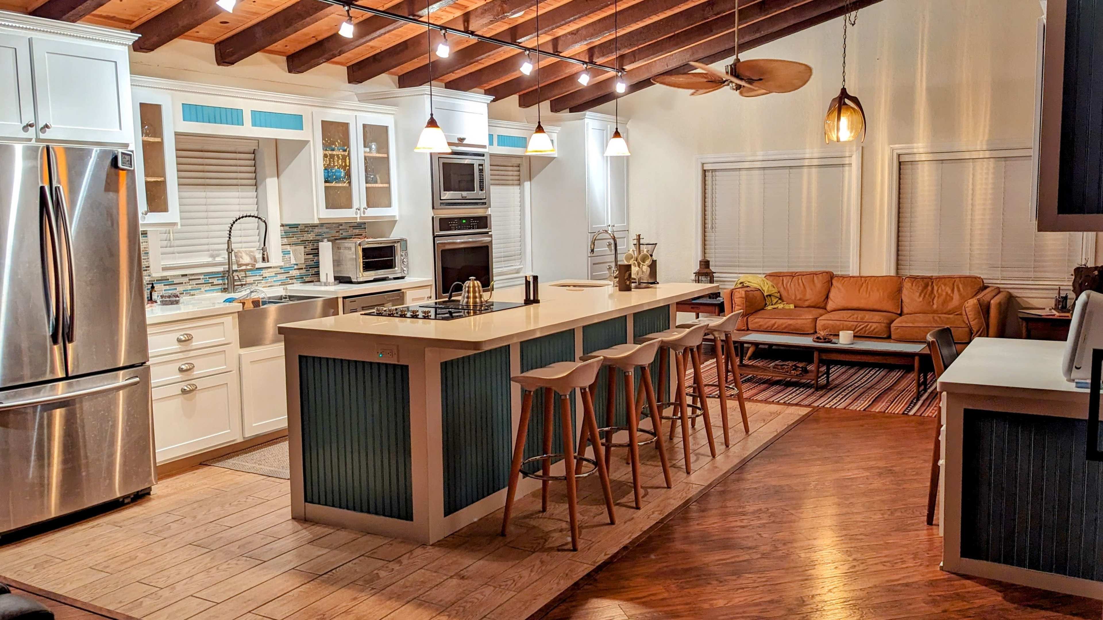 The image shows a modern kitchen with a central island and seating, adjacent to a living area featuring a leather couch and wooden flooring.
