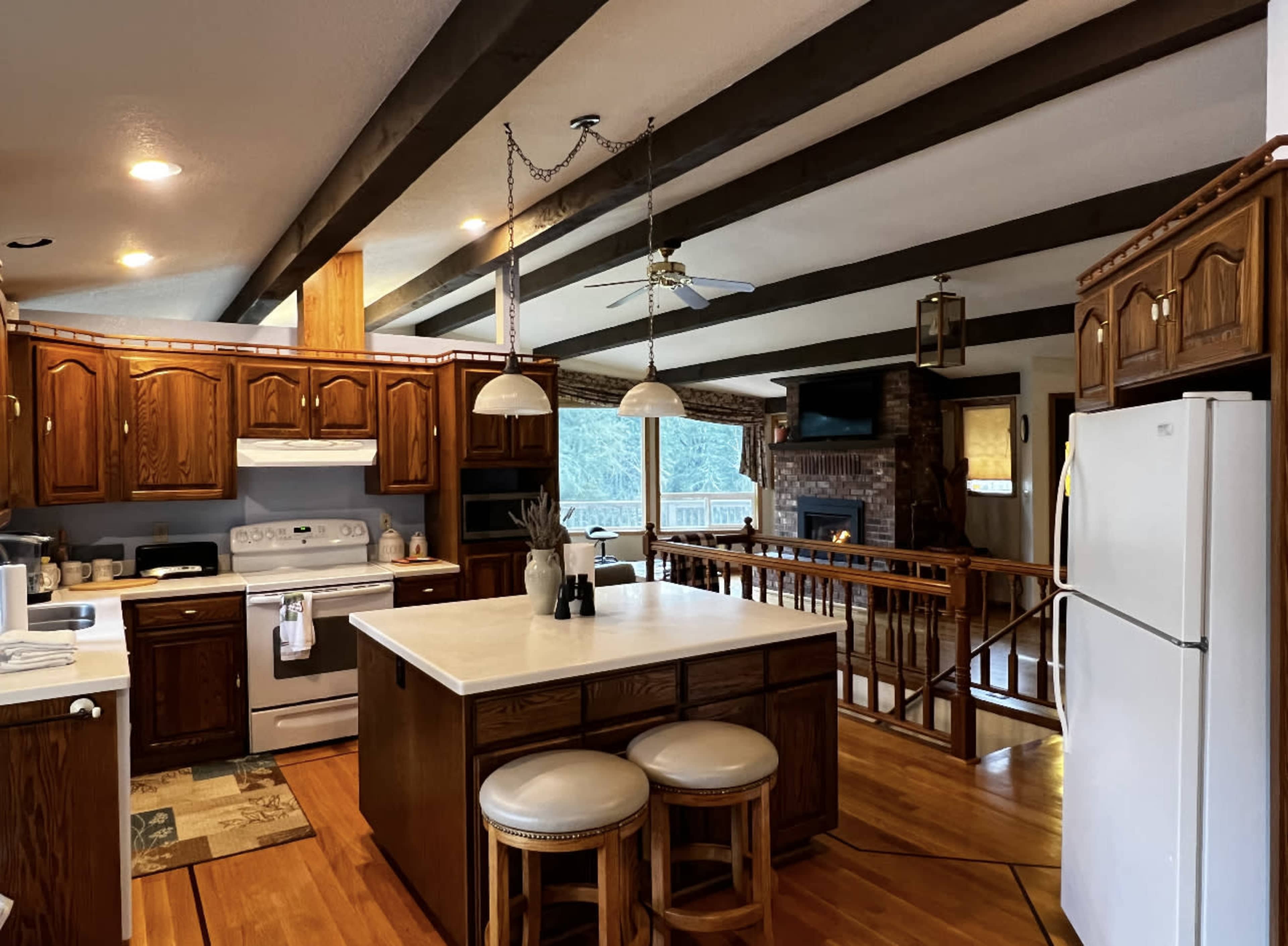 The kitchen features wooden cabinets, a center island with bar stools, and an open layout that connects to a living area with a fireplace and large windows.