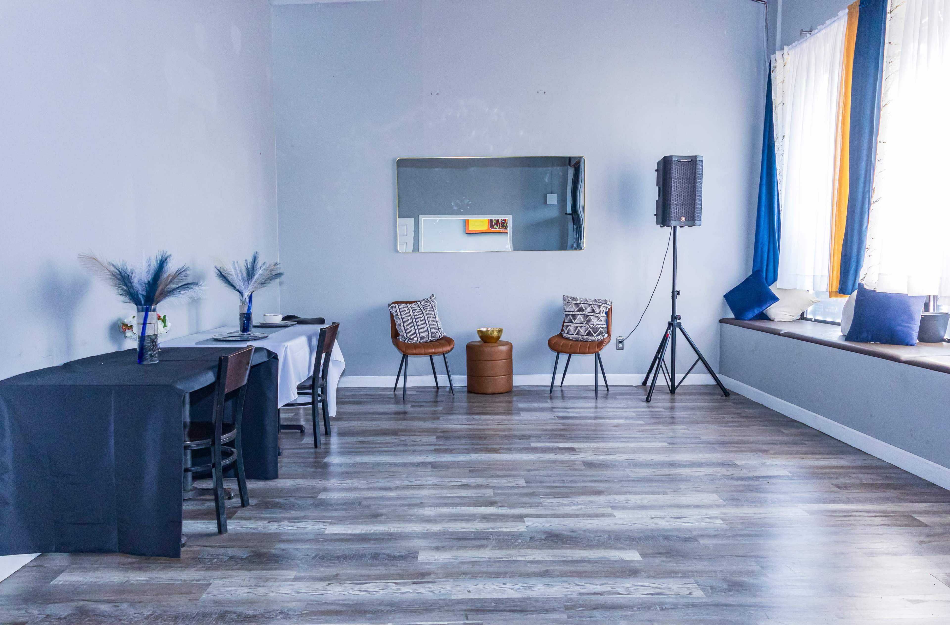 The image shows a minimalist room with gray walls, featuring a table, two chairs, a mirror, a speaker, and a window with blue curtains.