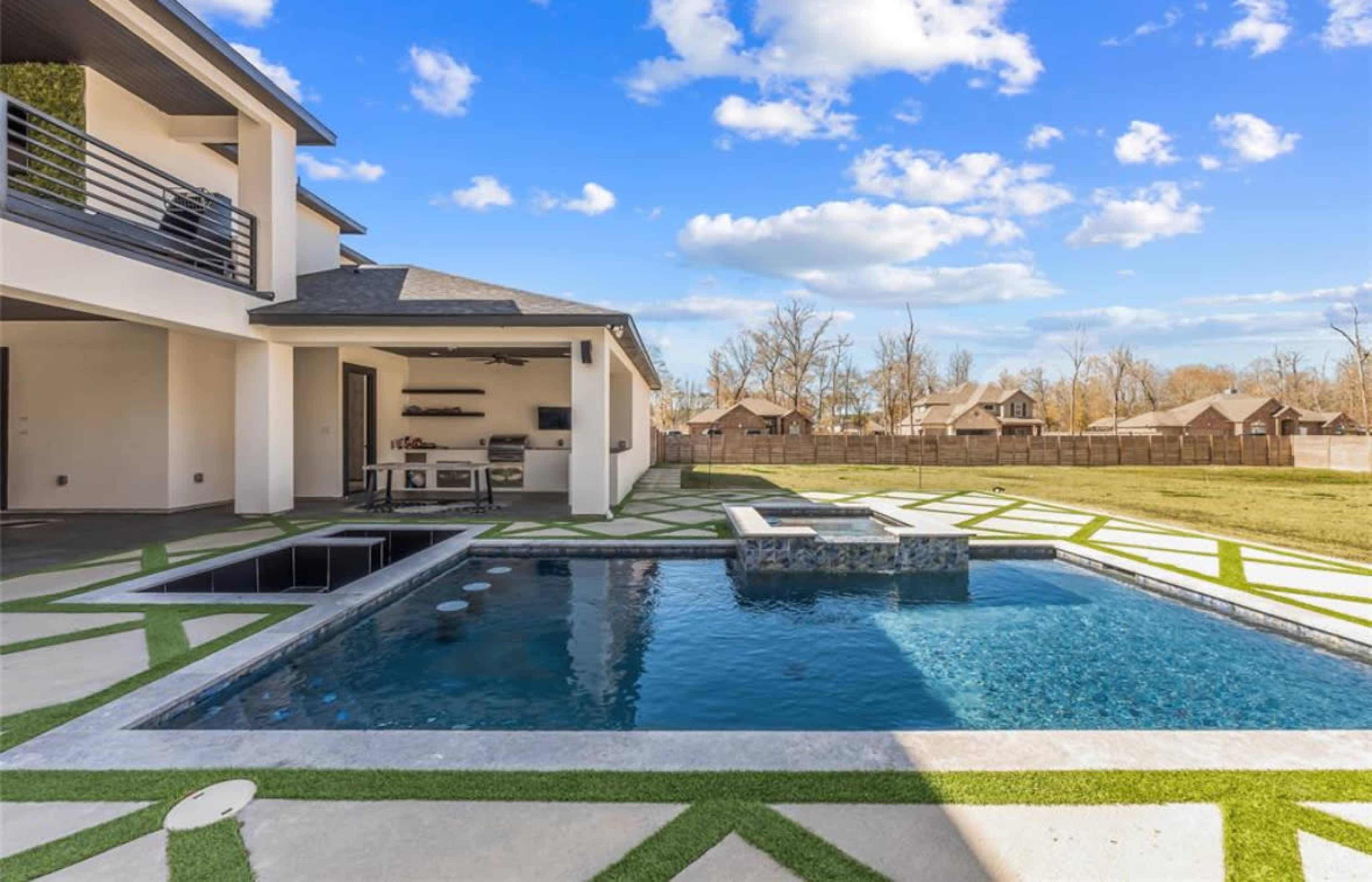 The image shows a modern backyard featuring a rectangular swimming pool with a spa and surrounding green grass landscaping, set against a backdrop of several houses.