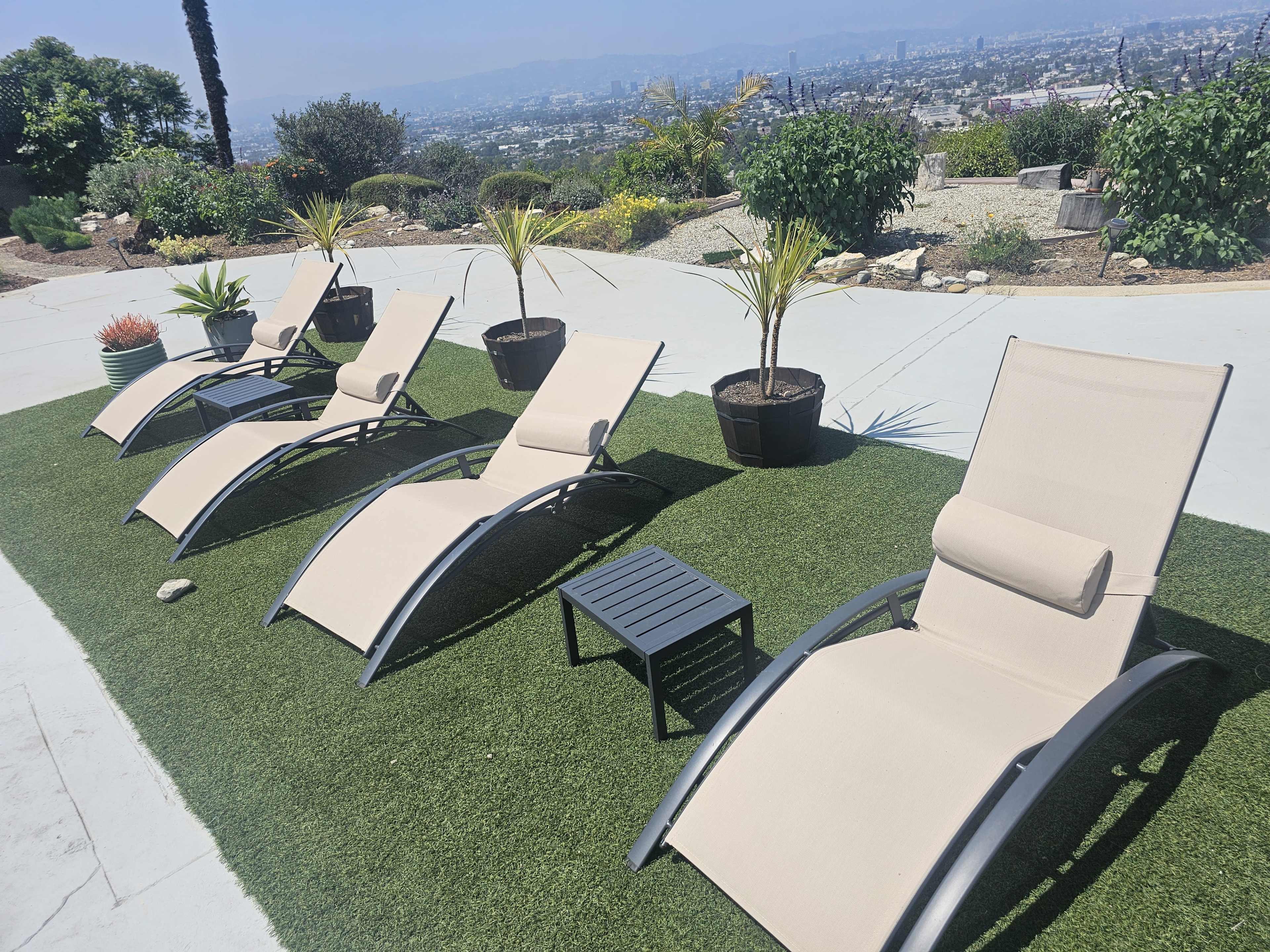 Four lounger chairs are arranged on a grassy area, accompanied by small tables and potted plants, with a view of a city landscape in the background.