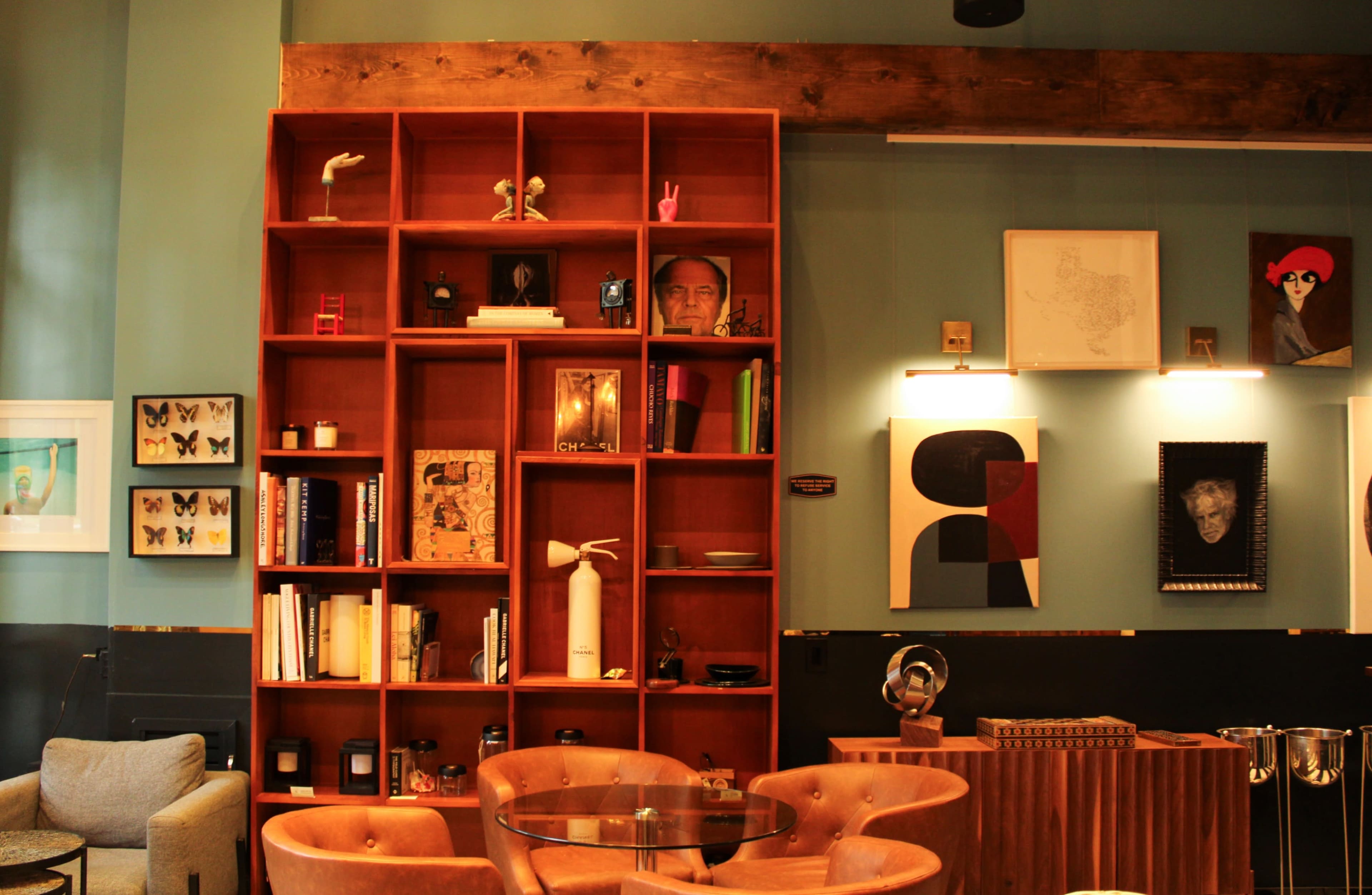 A cozy interior of a café or lounge, featuring a wooden shelf filled with books and decorative items, surrounded by modern furniture and art pieces on the walls.