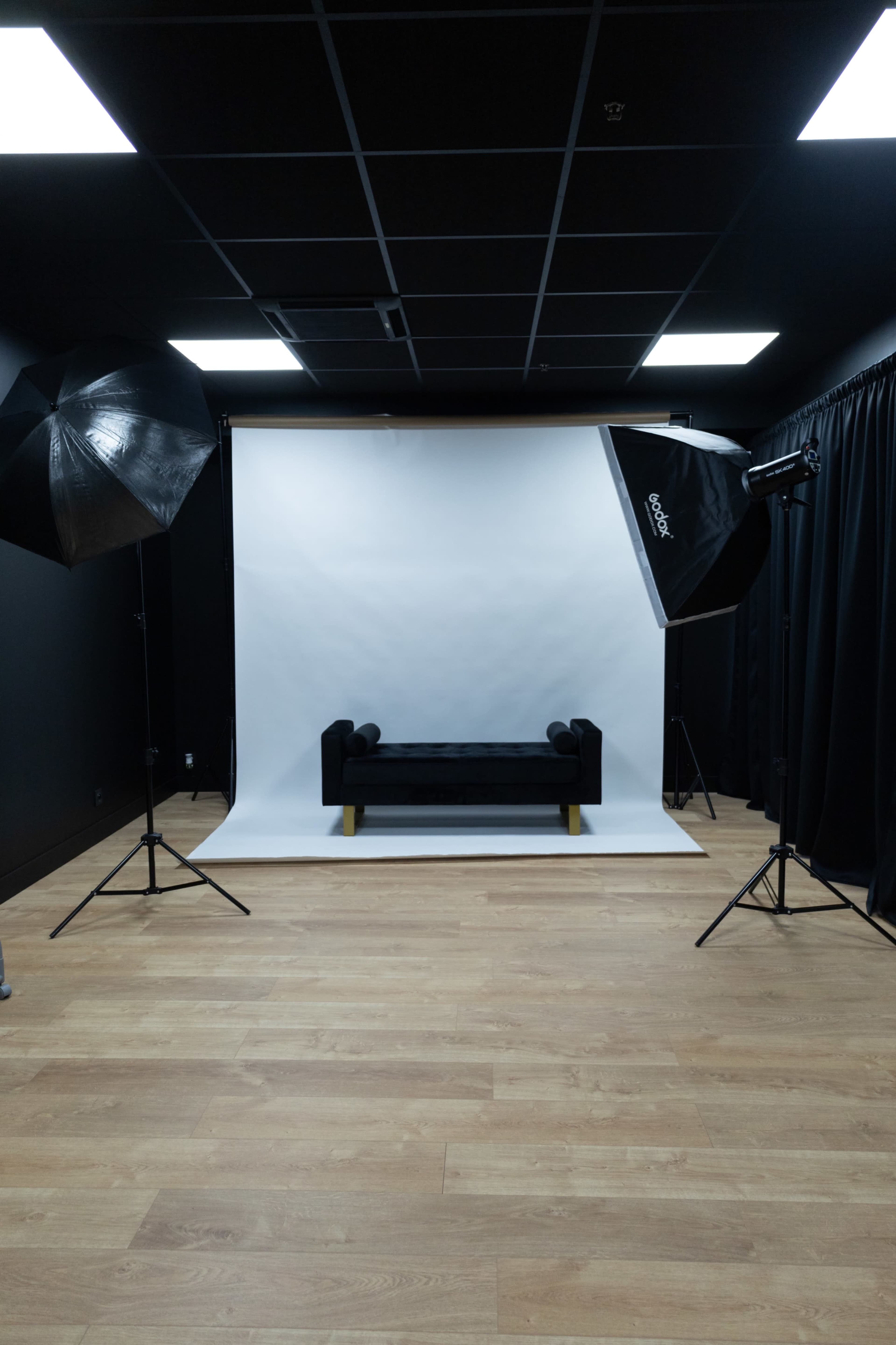 A photo studio is set up with a black couch centered on a white backdrop, flanked by two large softbox lights.