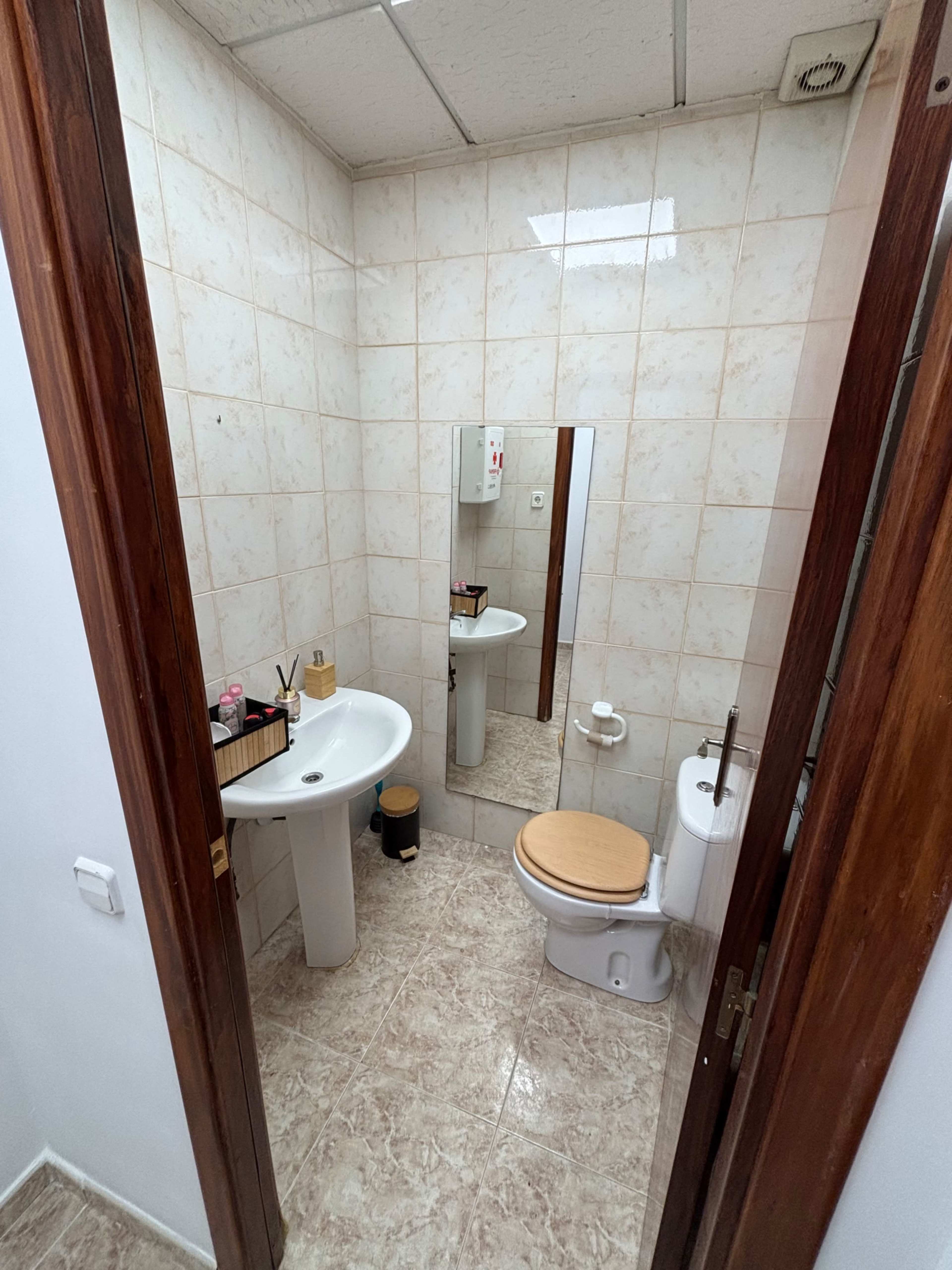 The image shows a small bathroom containing a sink, a toilet, and a mirror on the wall, all surrounded by tiled walls and a tiled floor.