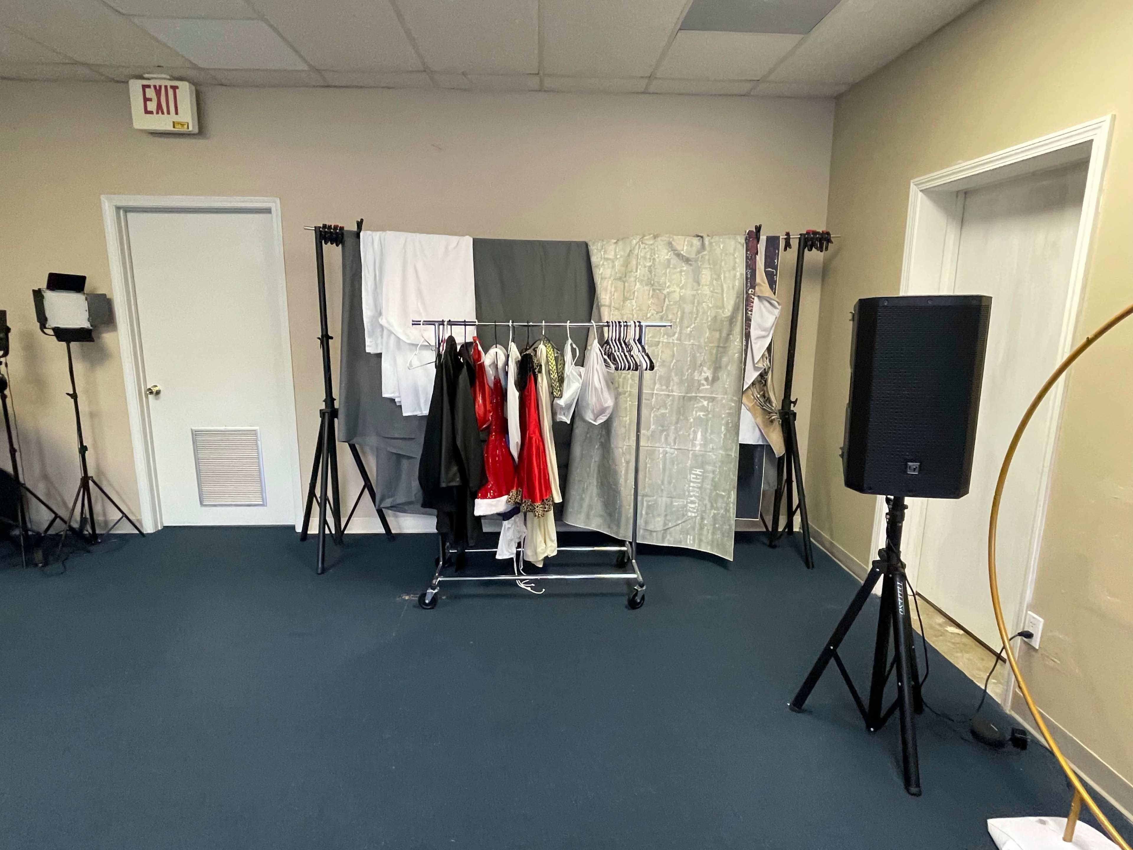 A clothing rack with various garments hung on it, positioned against a backdrop of draped fabric in a room with beige walls and blue carpet.