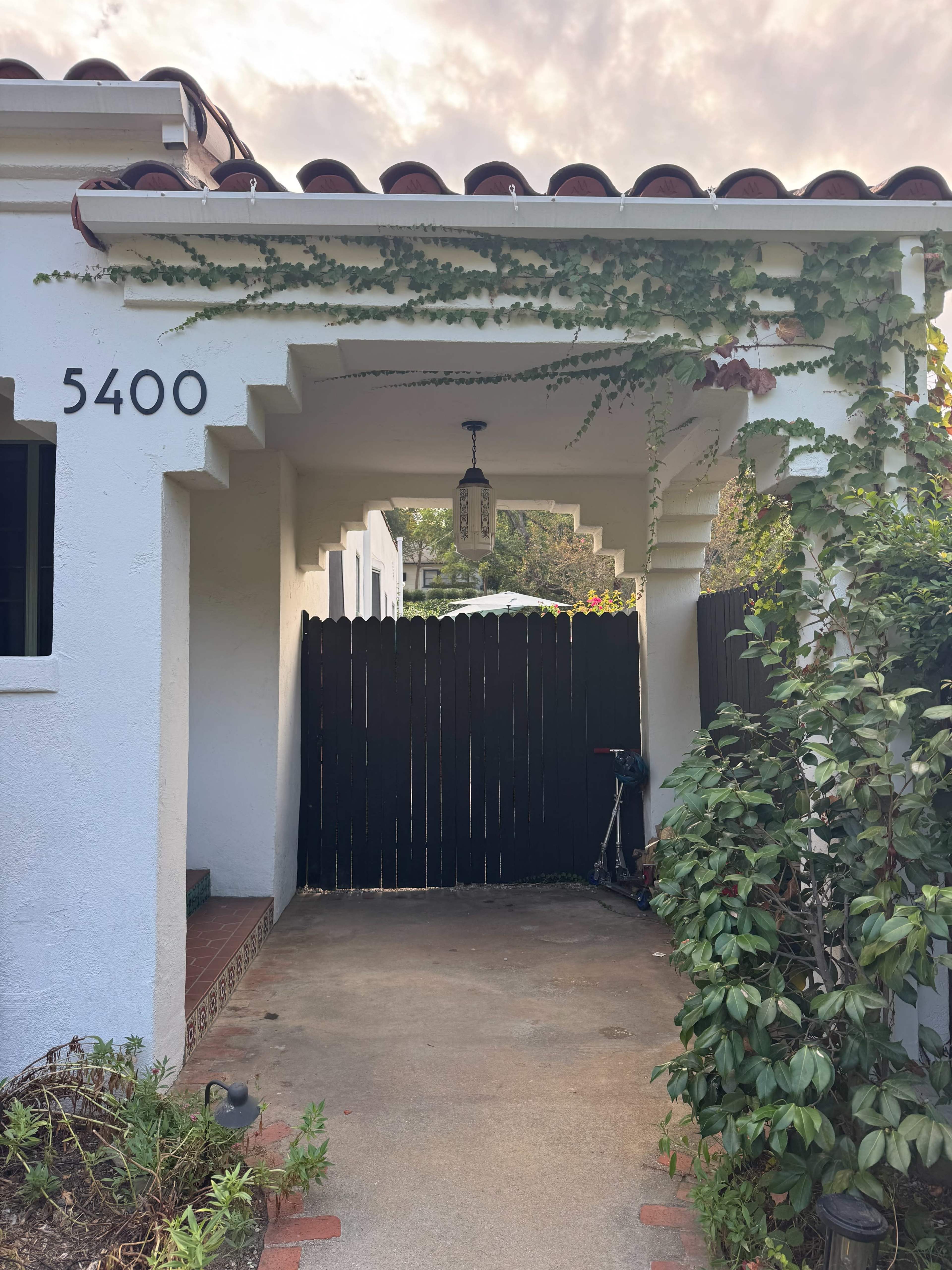 The image shows an entrance to a house with a tiled roof, white walls, a hanging light fixture, and a wooden fence in the background.