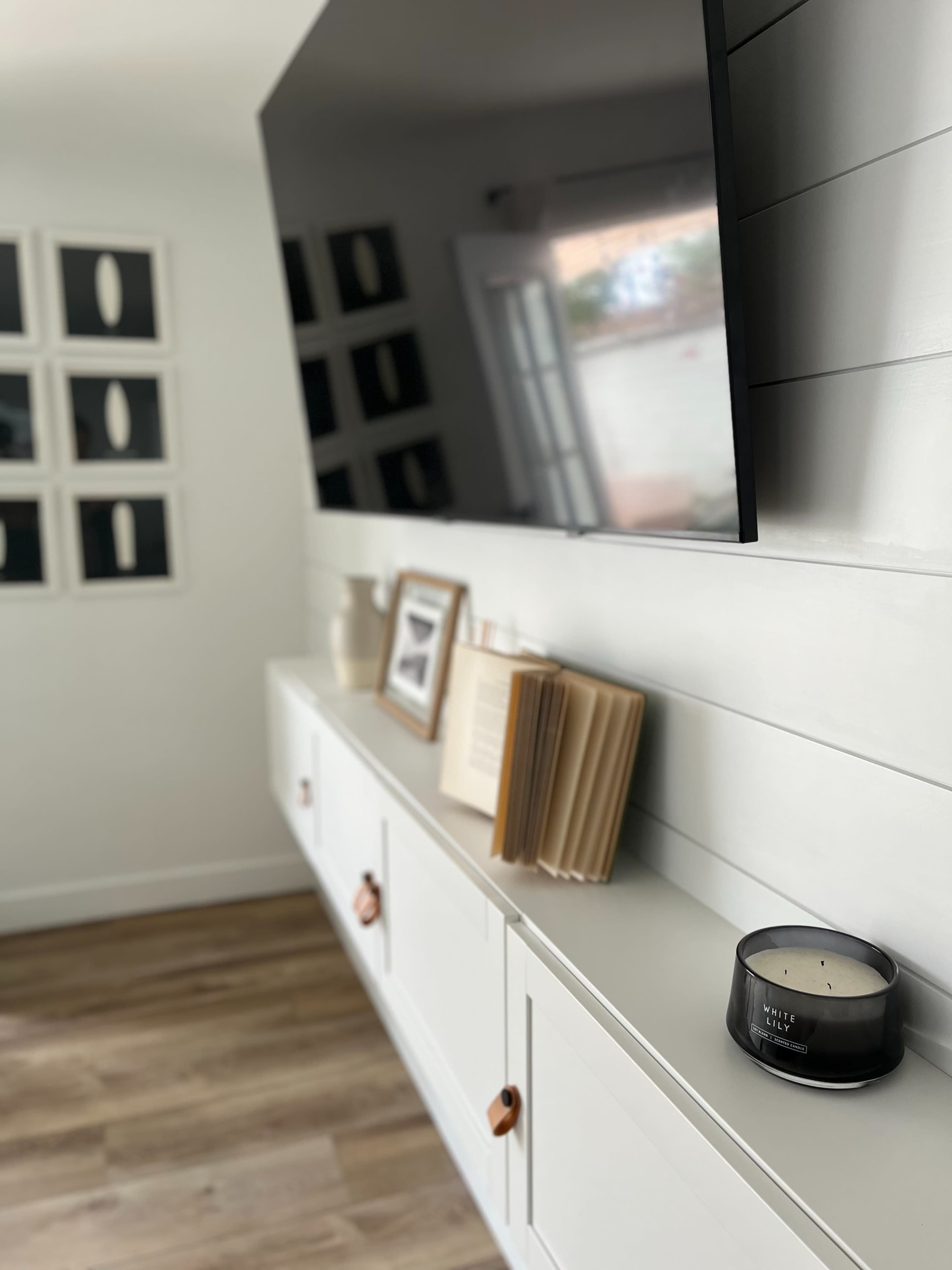 A modern living room features a wall-mounted TV above a sleek white cabinet with wooden handles, accompanied by framed artwork and a candle on the surface.