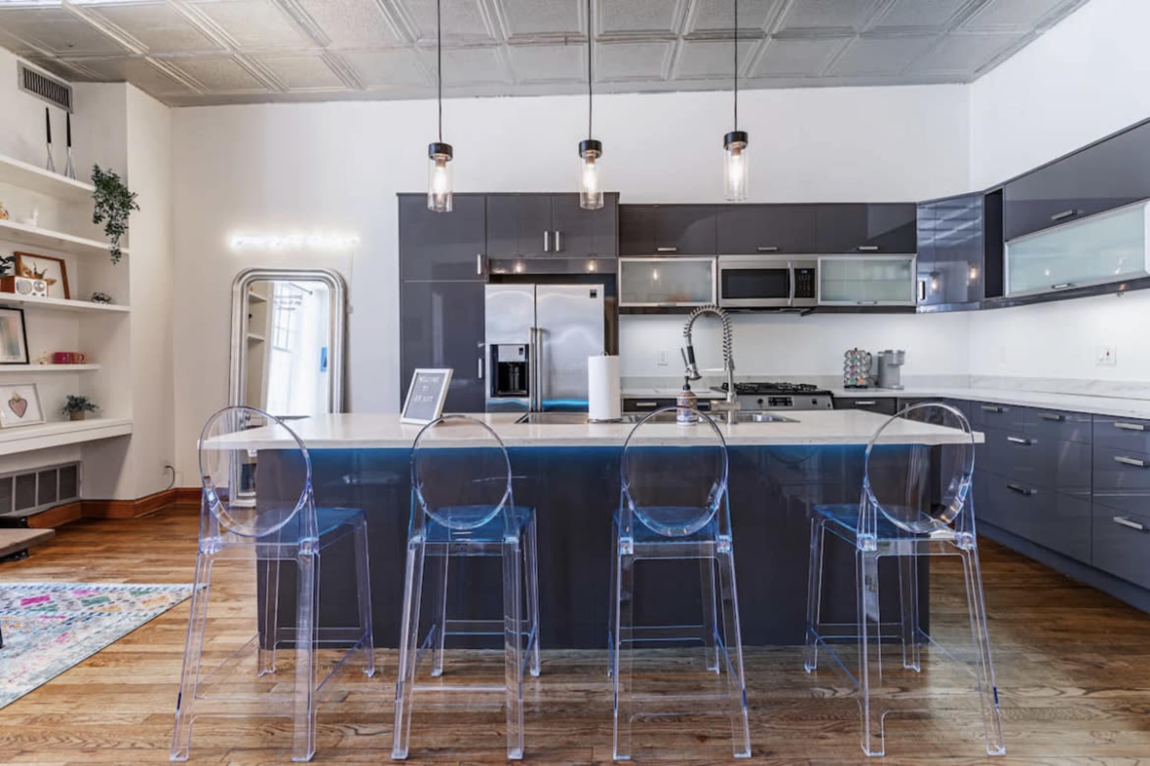 A modern kitchen features a sleek island with four clear acrylic stools, stainless steel appliances, and contemporary cabinetry.