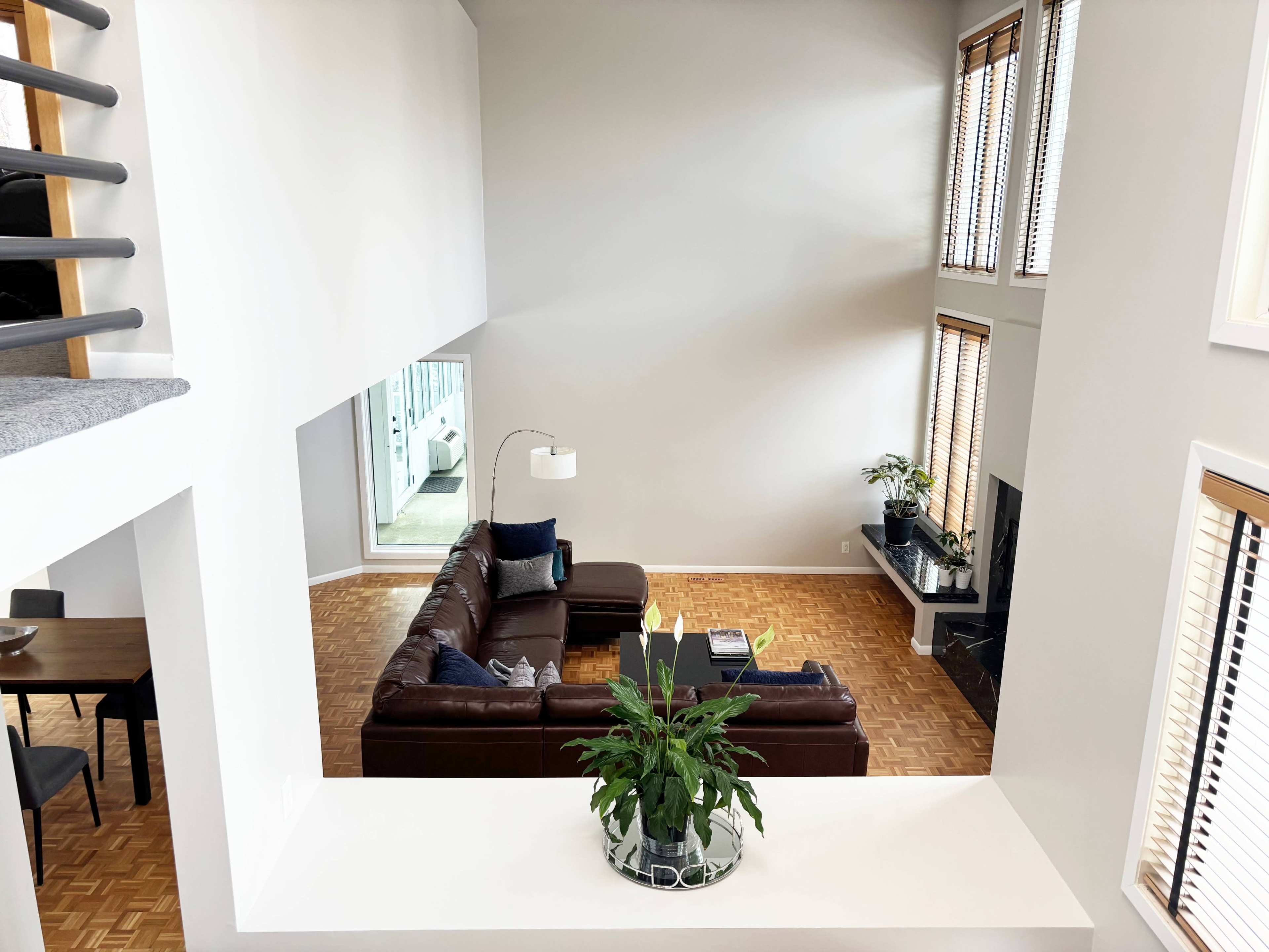 The image shows a spacious living room with large windows, a sectional sofa, and a potted plant in the foreground.
