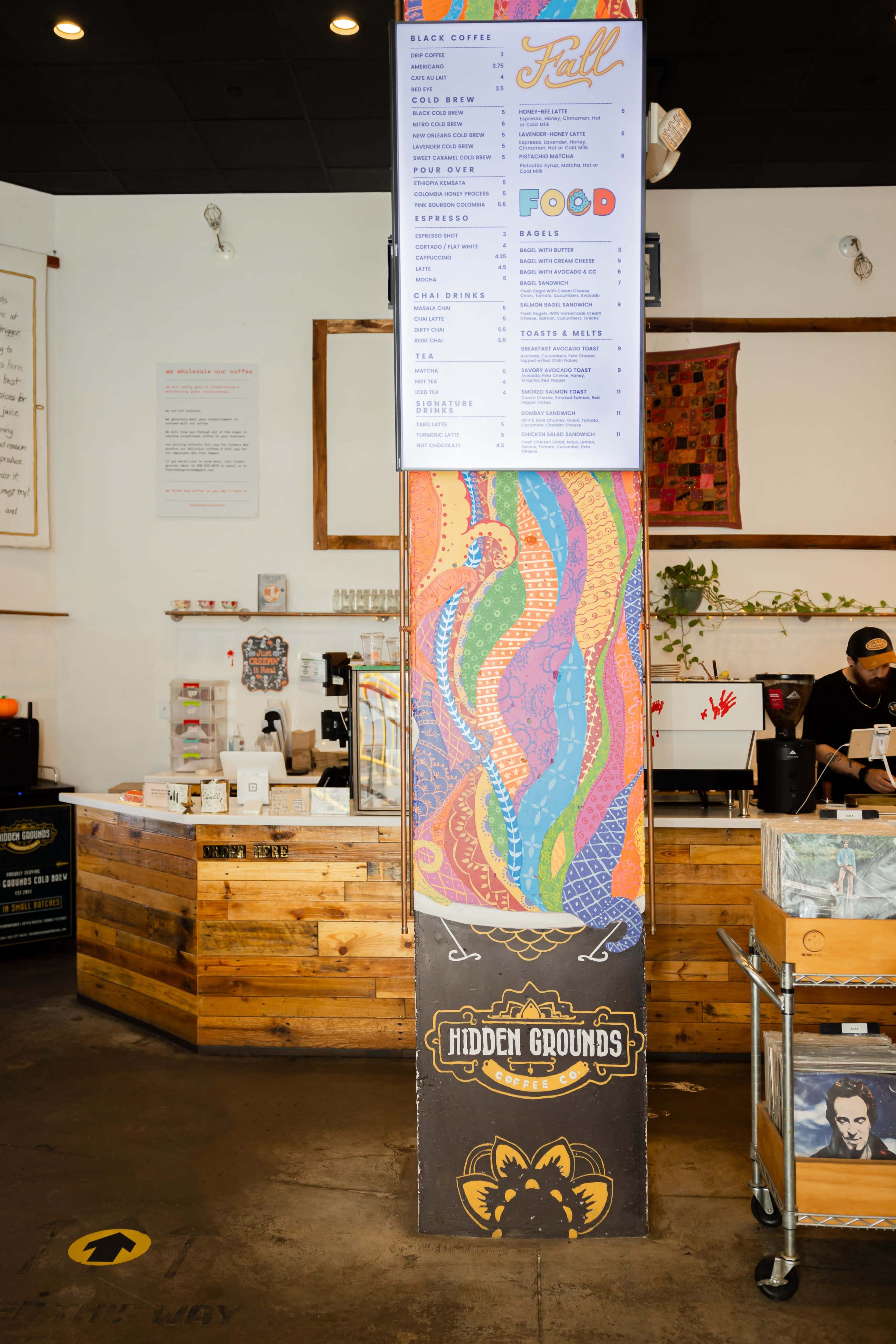 The interior of a coffee shop featuring a colorful mural on a central pillar, a wooden counter, and a menu displayed above.