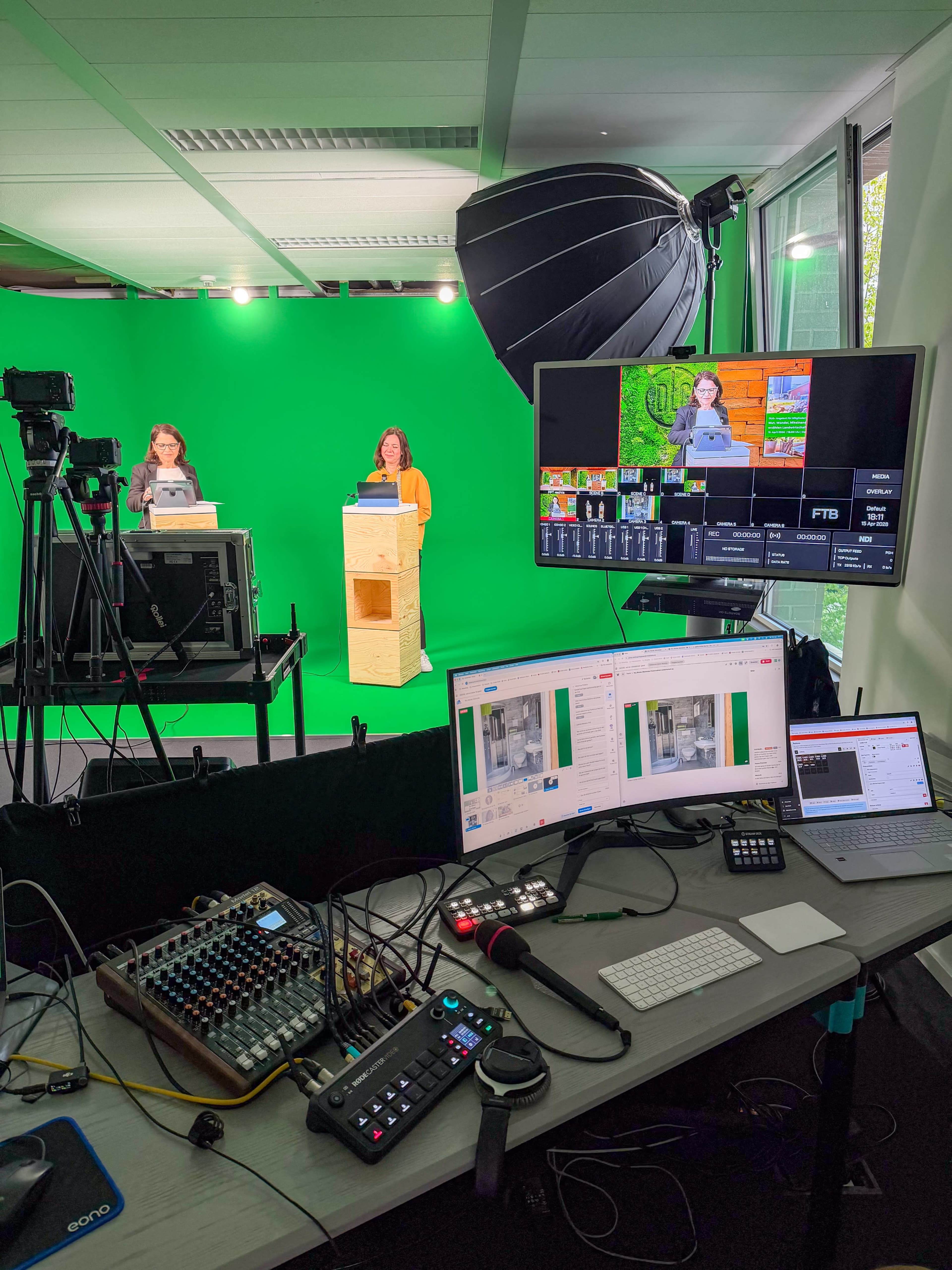 The image shows a green screen studio setup with two women presenting at a podium, surrounded by cameras, audio equipment, and a computer monitor displaying video feeds.