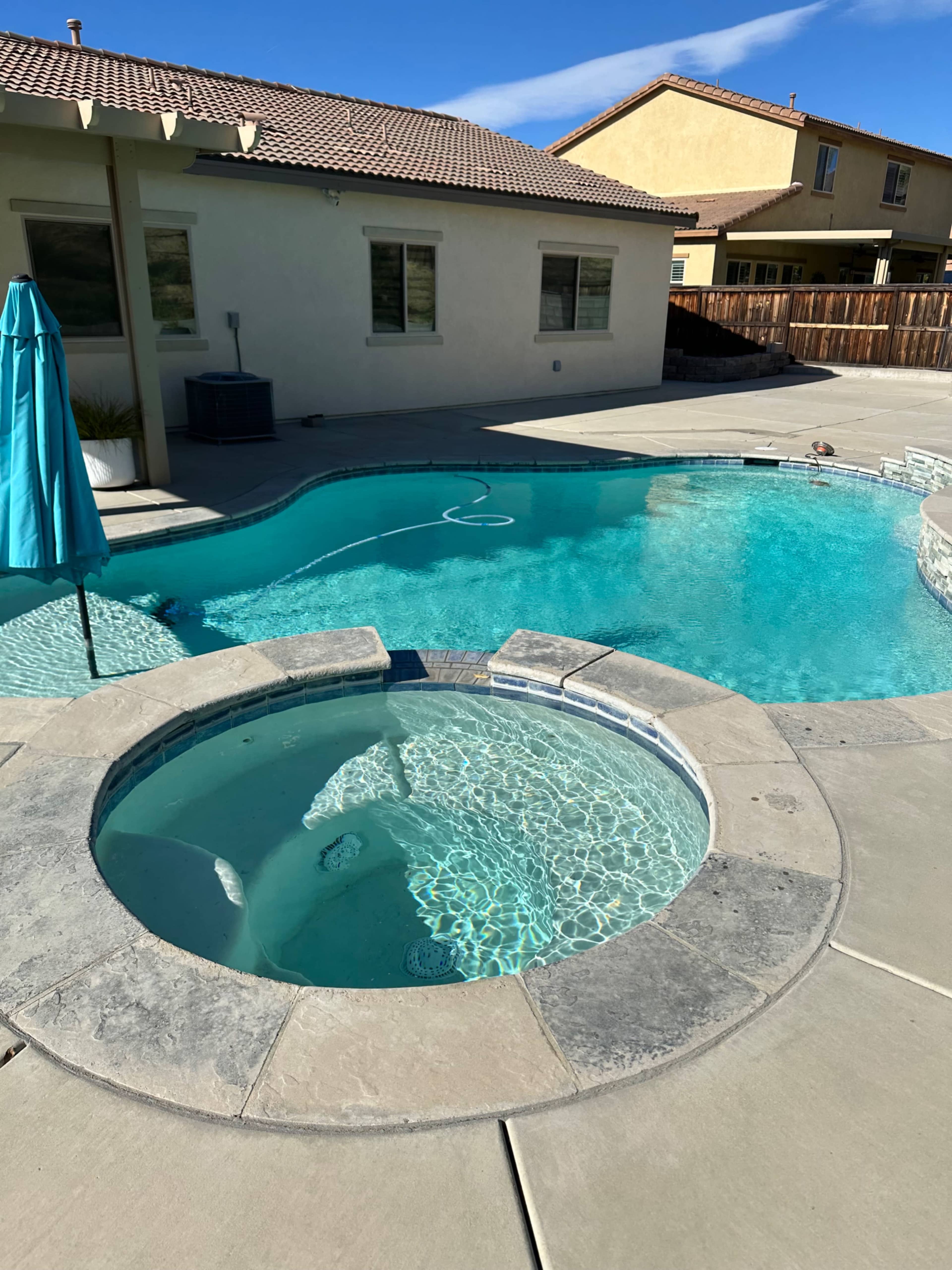 The image shows a residential backyard featuring a clear swimming pool with a separate hot tub, surrounded by concrete paving and a patio area.