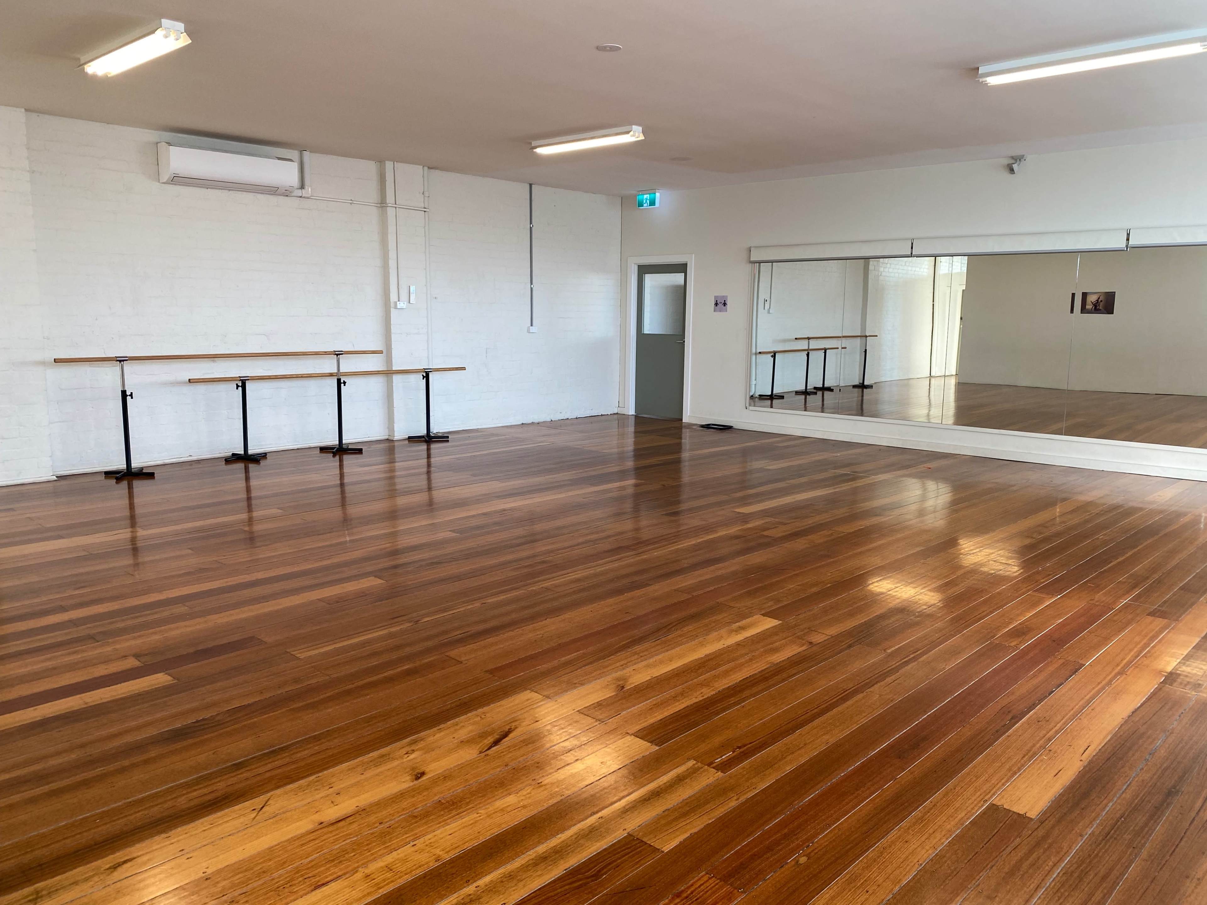 The image shows a spacious dance studio with polished wooden floors, mirrored walls, and several wooden barres along one side.