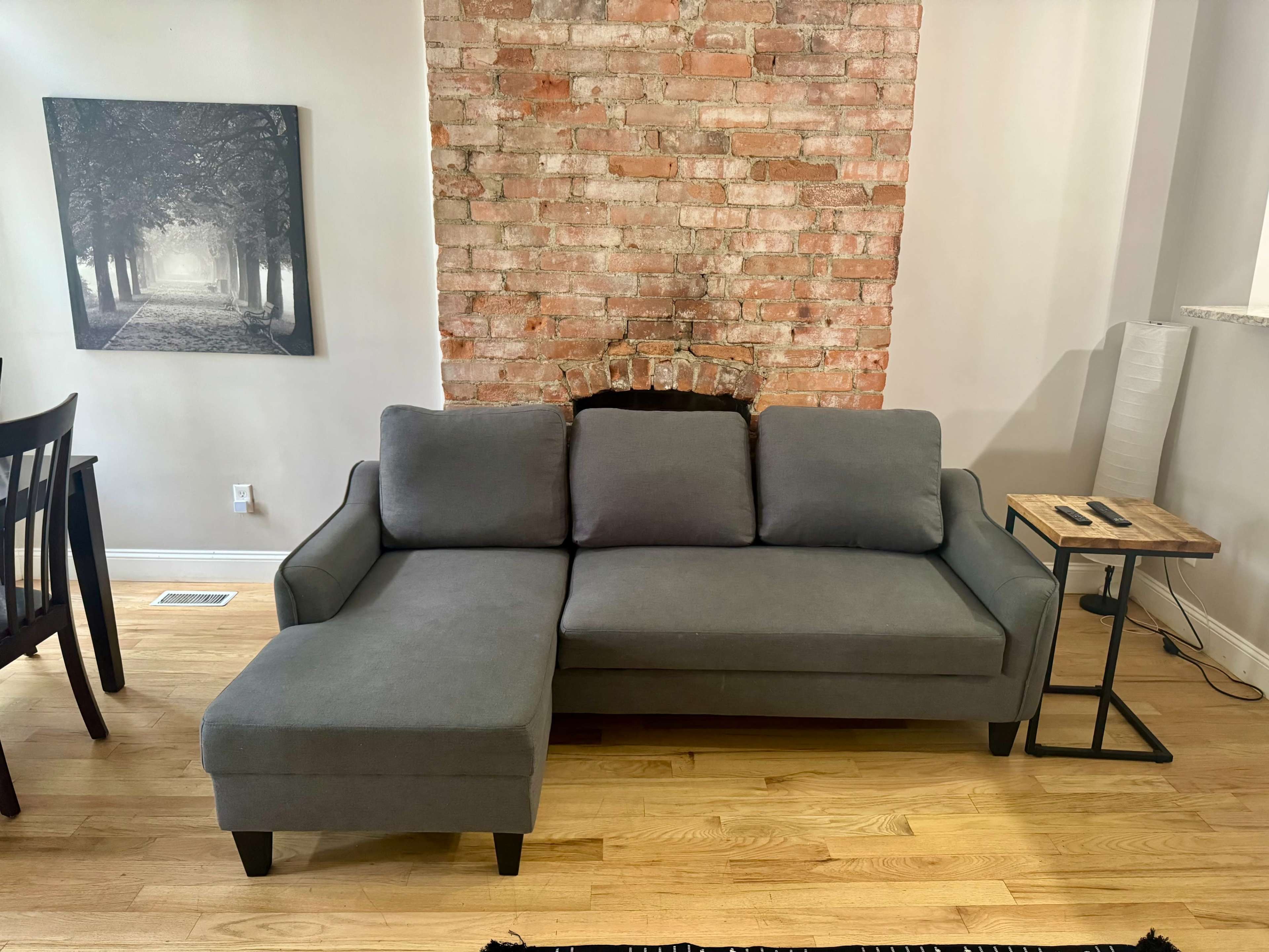 The image shows a gray sectional sofa positioned against a brick wall, with wooden flooring and a small table nearby.