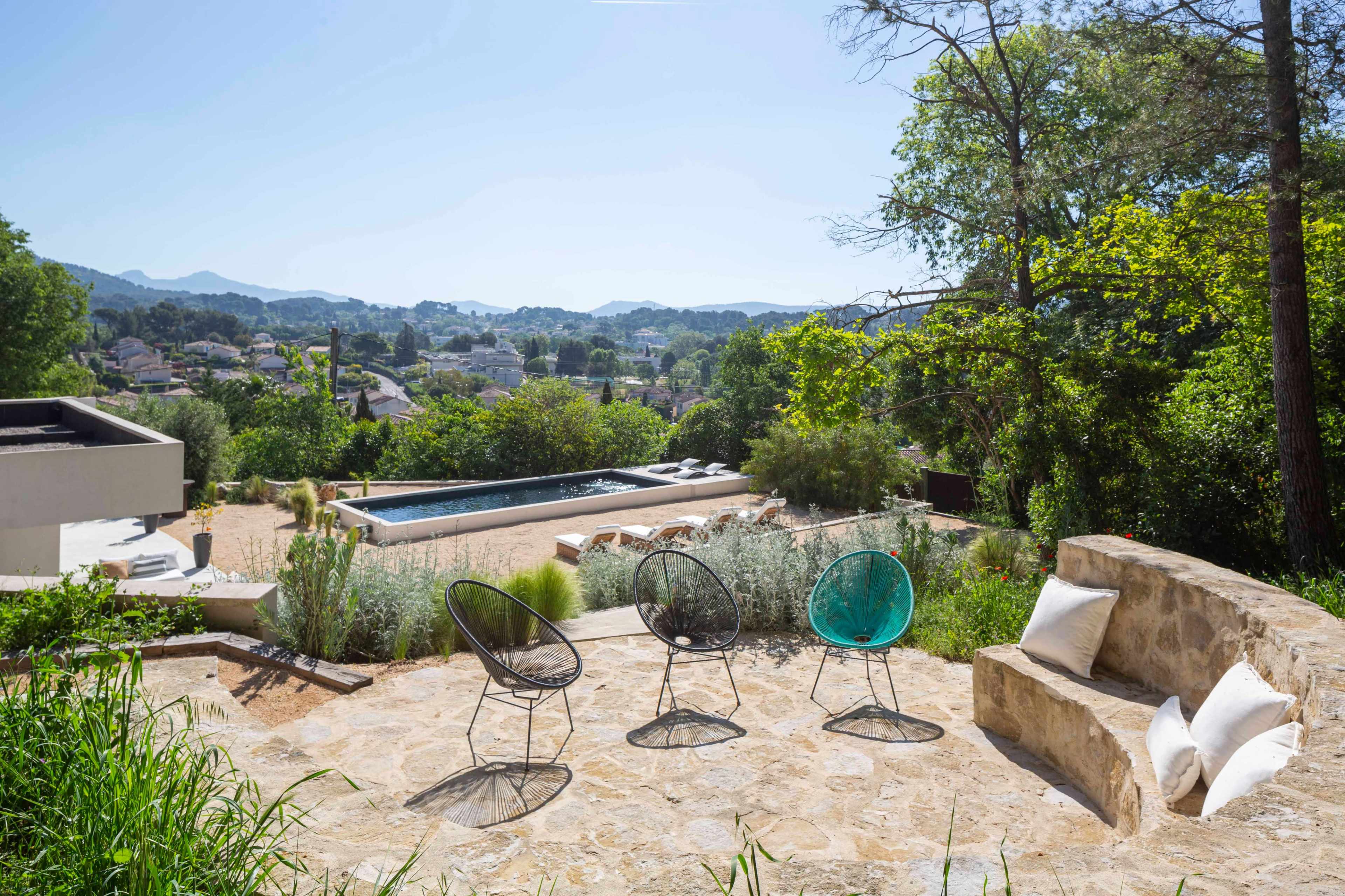 The image shows a scenic outdoor space with four chairs arranged on a stone patio overlooking a hillside, featuring a swimming pool and landscaped greenery.