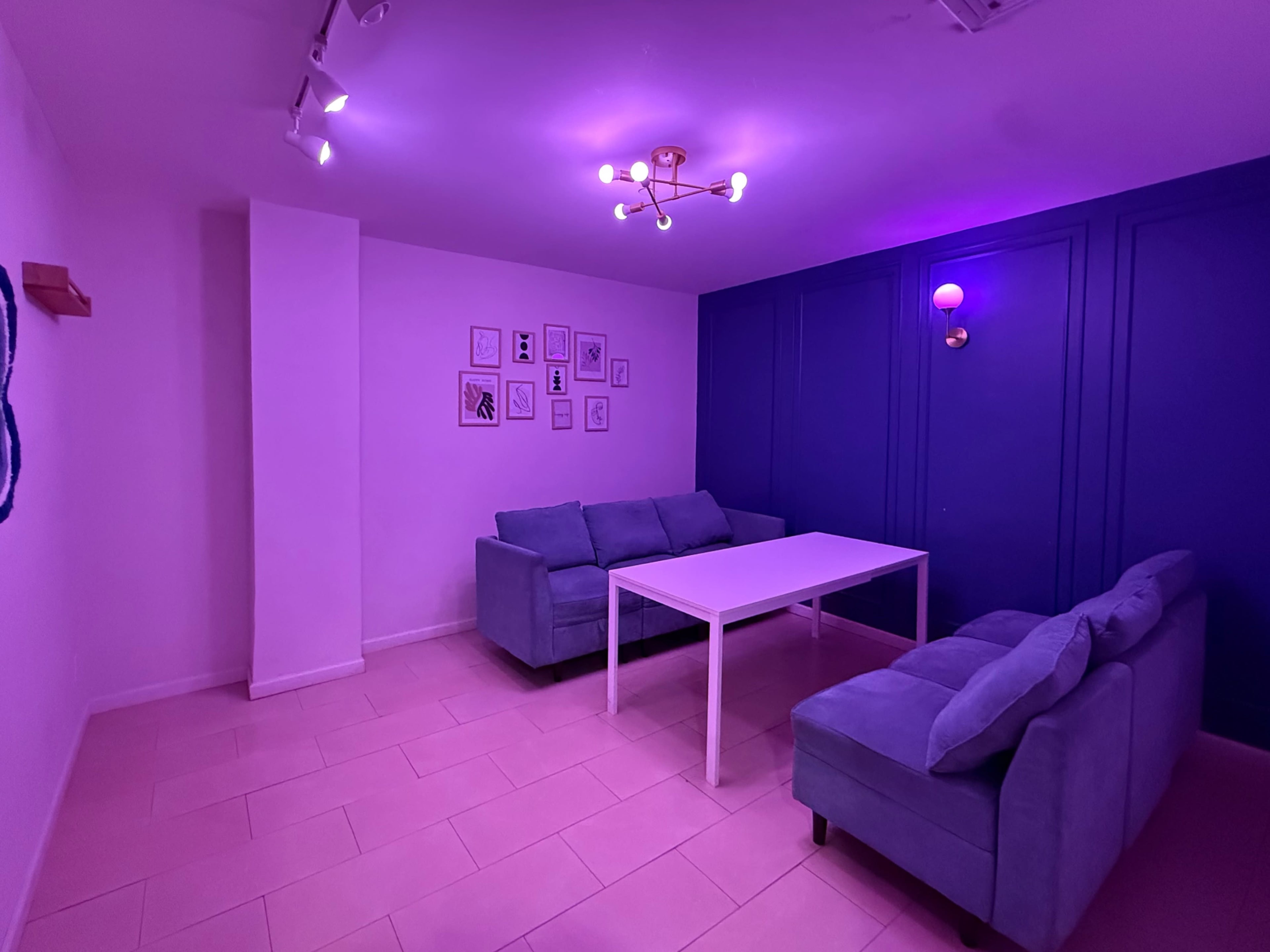 The room features a white table and two light blue sofas under purple lighting, with framed artwork on the wall.