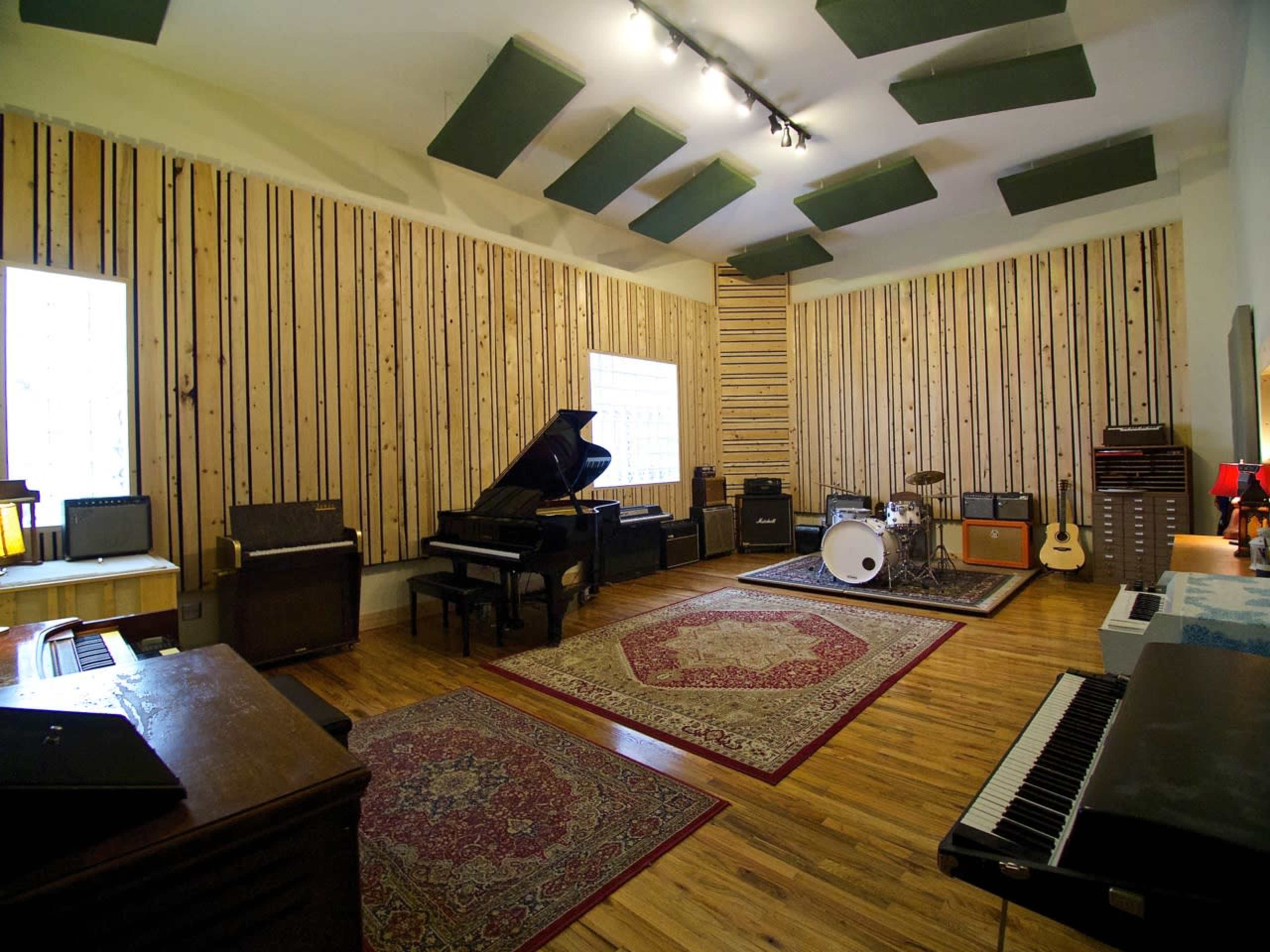A spacious music studio featuring a grand piano, a drum set, an electric guitar, and various amplifiers arranged around a patterned rug on a wooden floor.