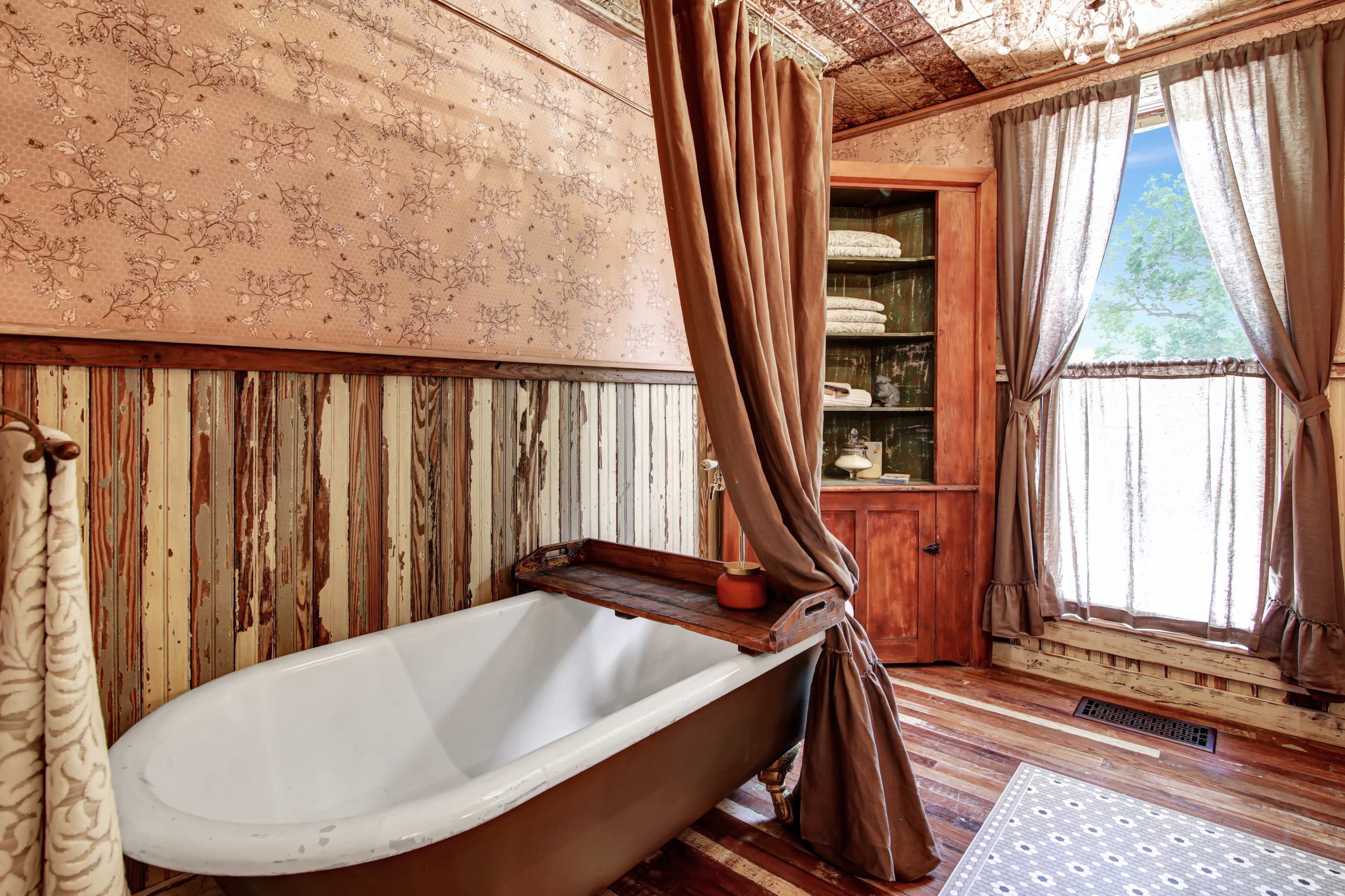 A vintage bathroom featuring a freestanding bathtub, wooden paneling, and large windows draped with curtains.