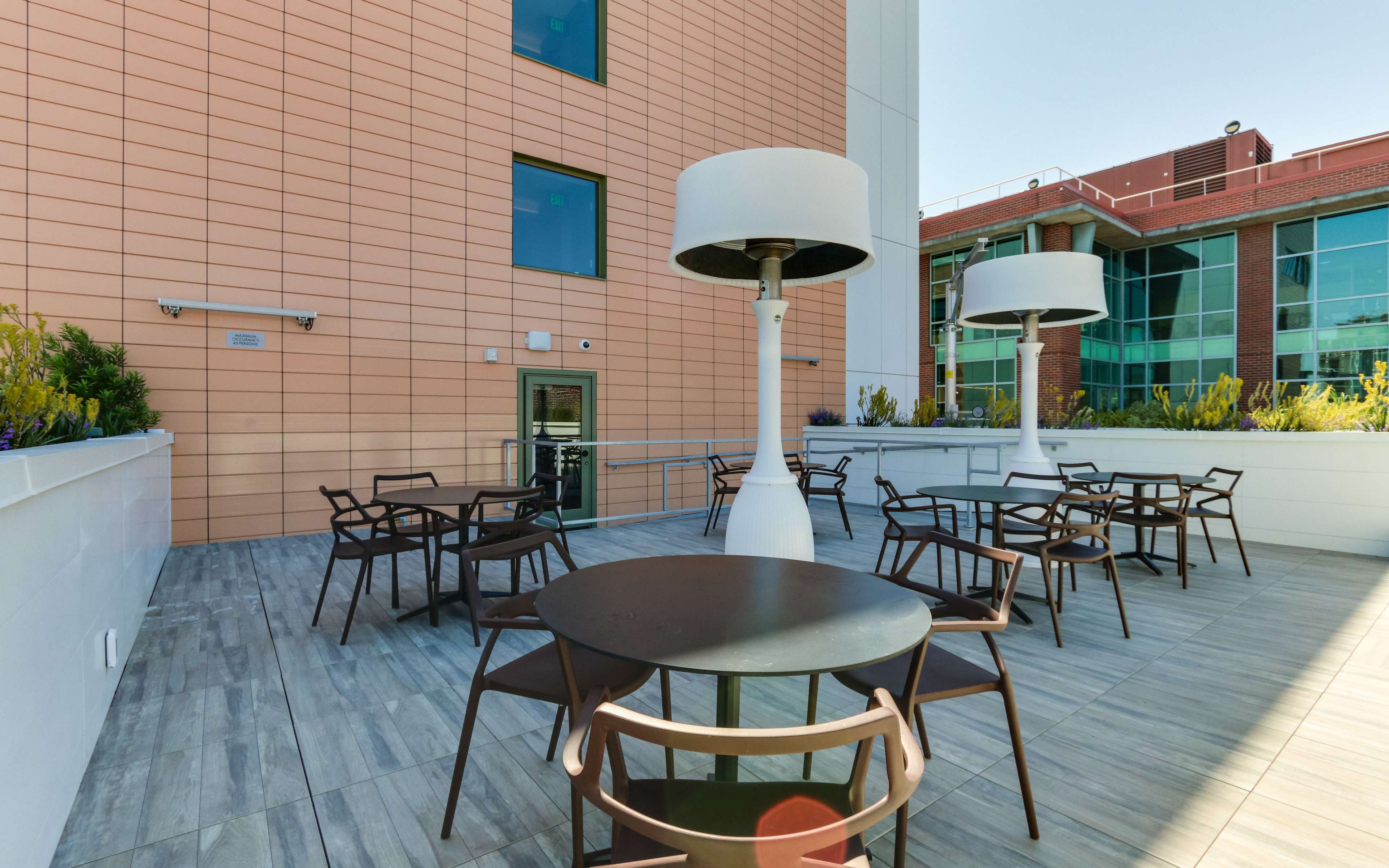 A terrace features several tables and chairs, accompanied by tall patio heaters and surrounded by green plants, adjacent to a modern building.