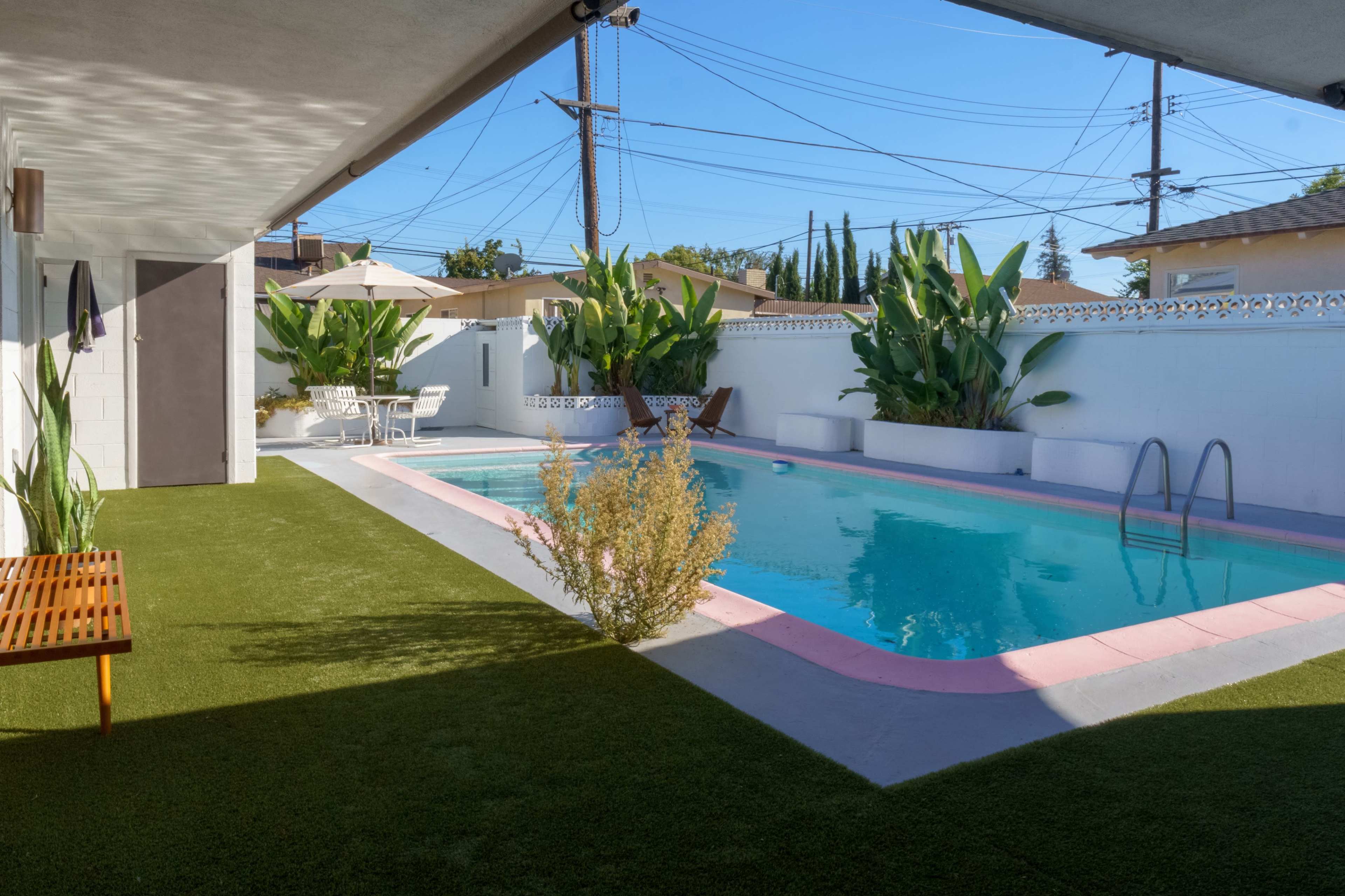 A swimming pool surrounded by lush green plants and a patio area with a table and chairs under an umbrella.