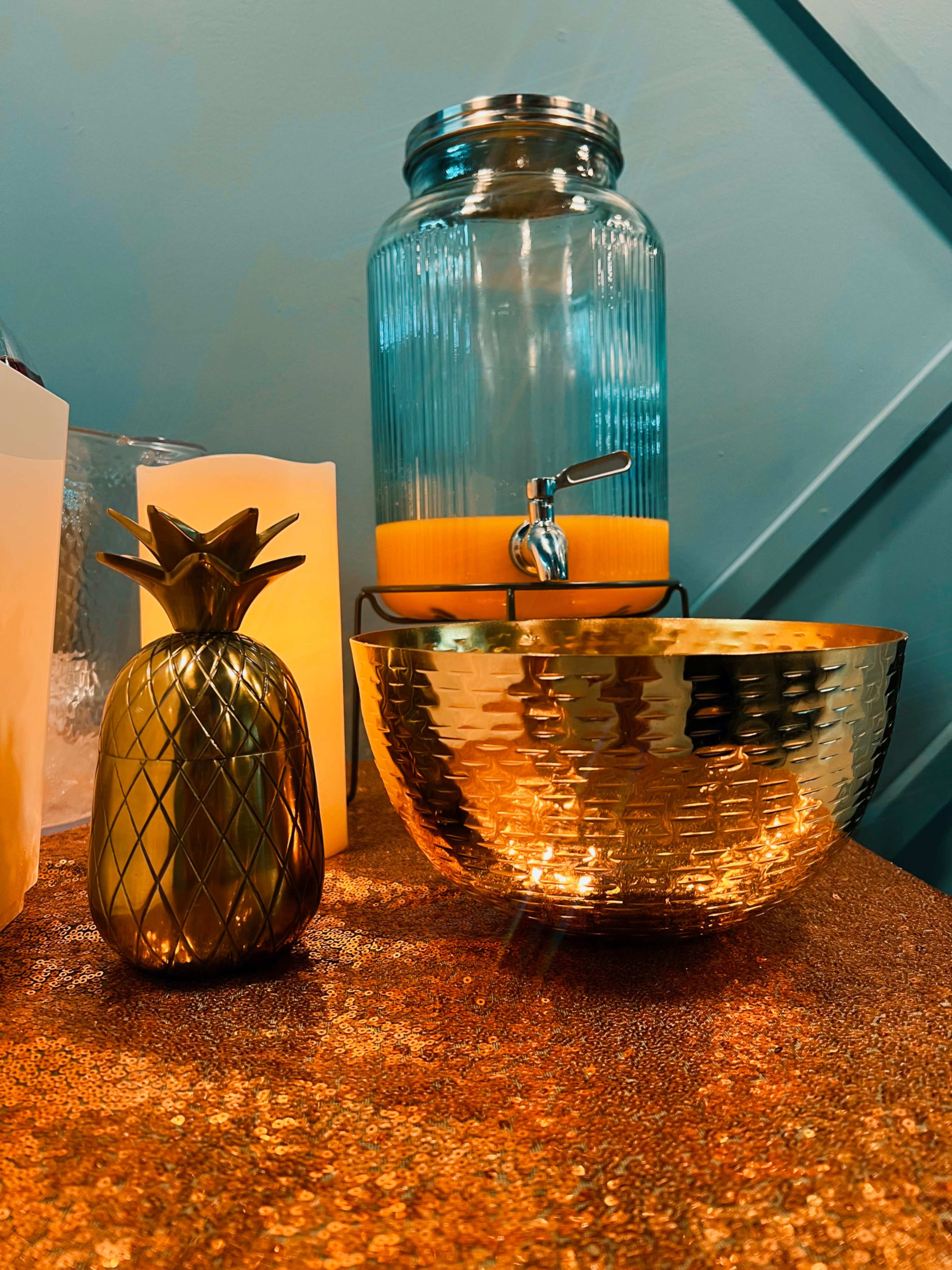 A golden pineapple figurine stands next to a glass jar filled with yellow liquid and a metallic bowl on a textured surface.