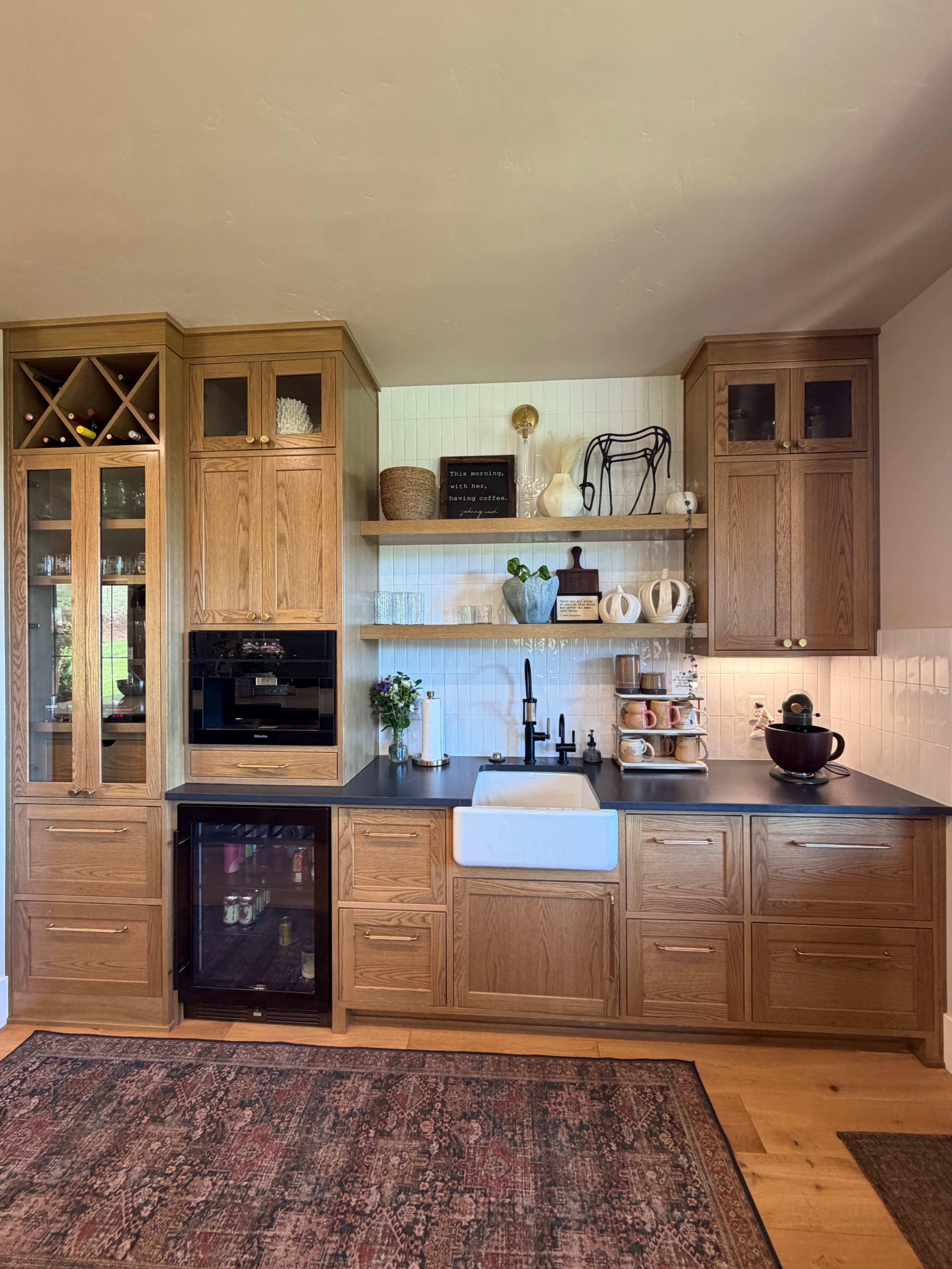 The image shows a modern kitchen with wooden cabinetry, a farmhouse sink, open shelving displaying dishware, and a wine rack integrated into the upper cabinets.