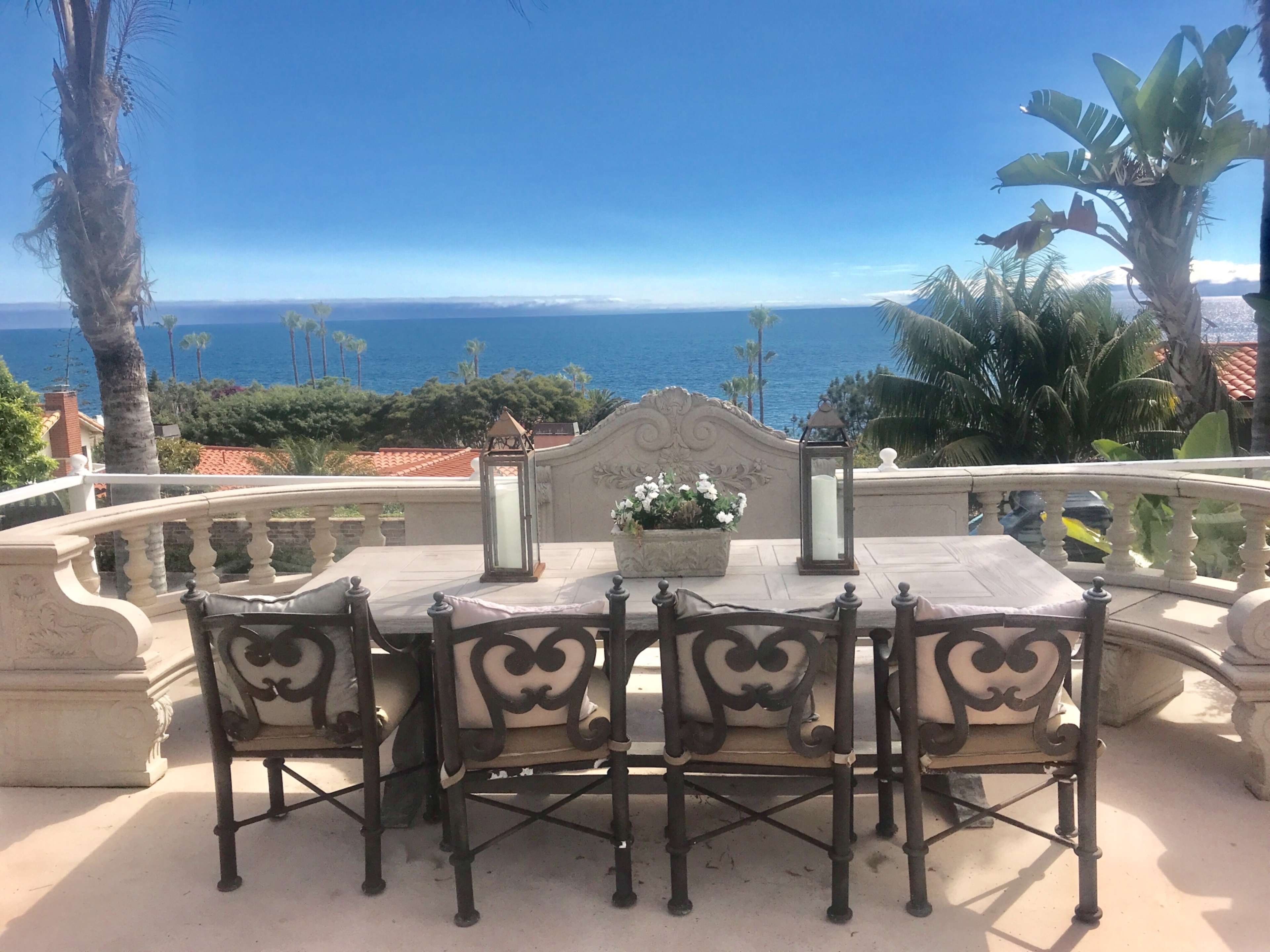 A dining table with six chairs is set on a terrace overlooking the ocean, framed by palm trees and plants.