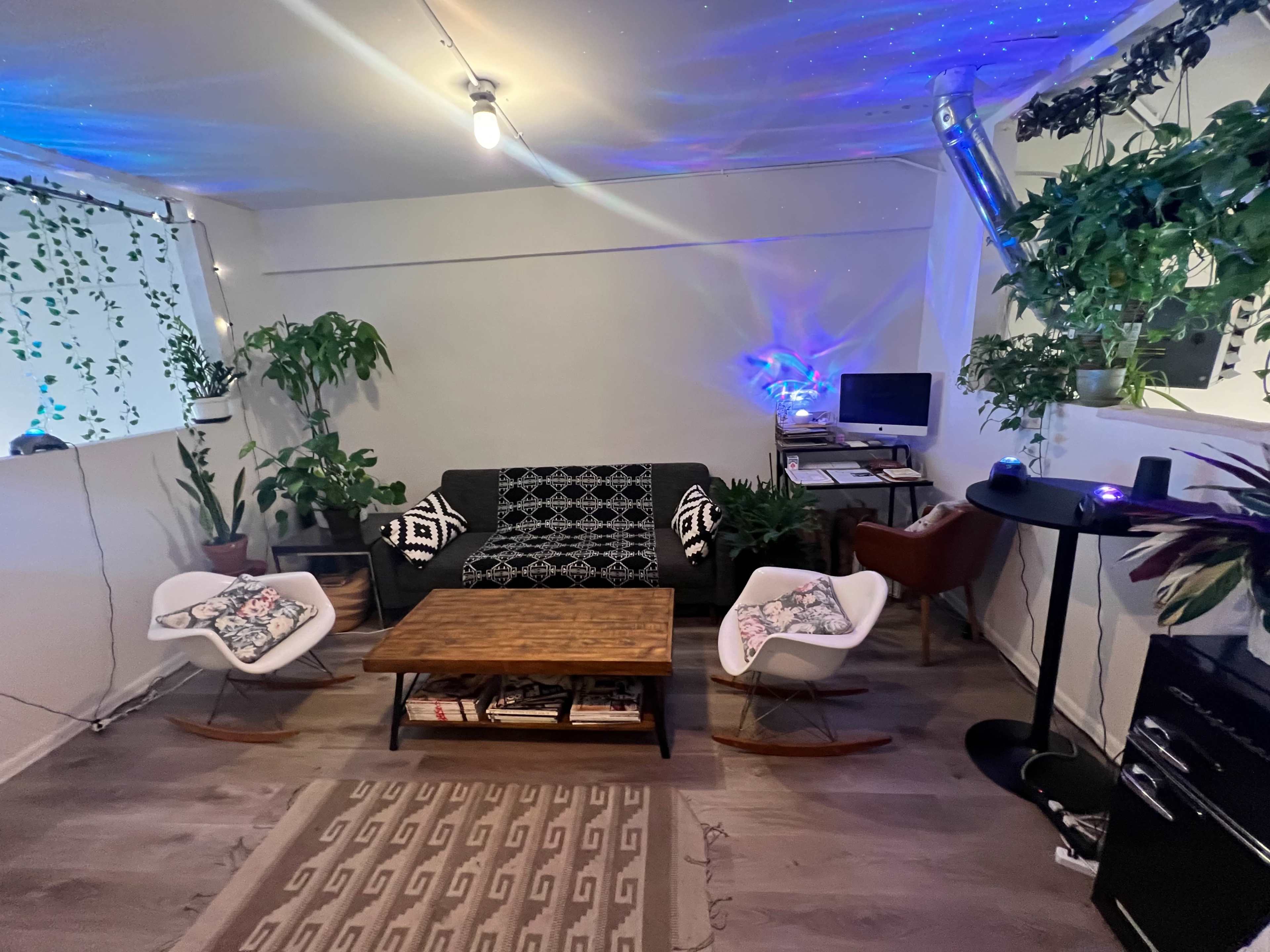 A cozy living space with a dark couch, two rocking chairs, a wooden coffee table, and various plants, illuminated by soft colored lighting.
