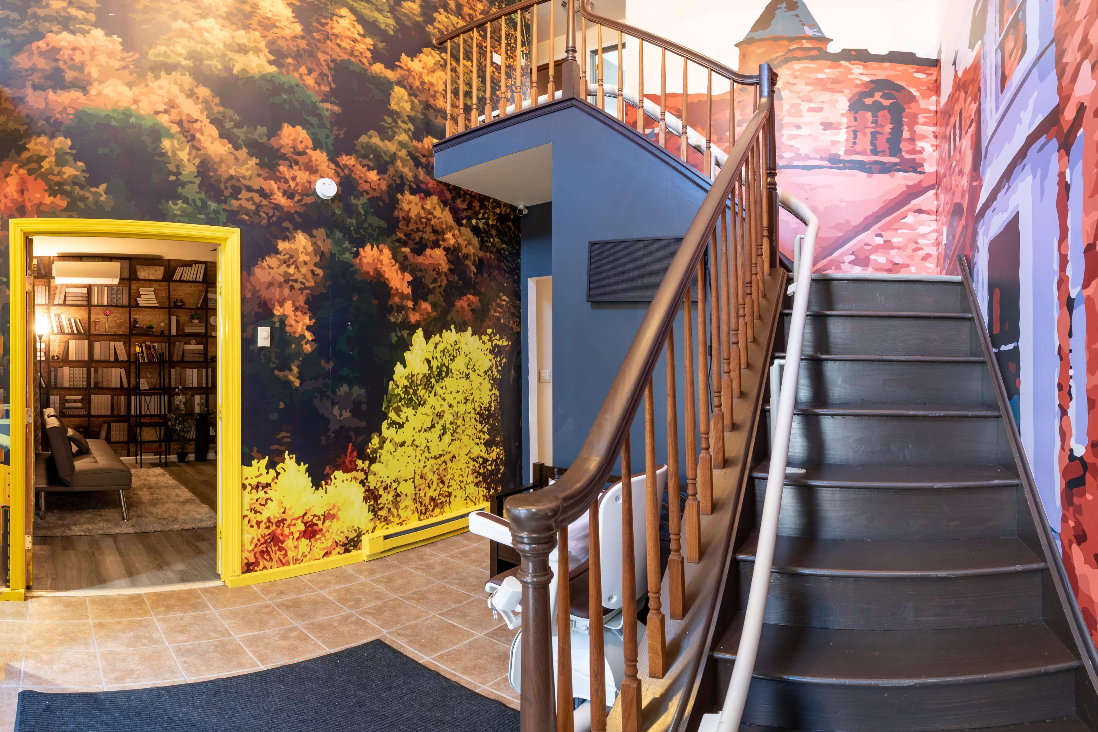 An entrance hall with a staircase, a vividly painted mural of autumn trees on the wall, and a bright yellow door leading to another room.