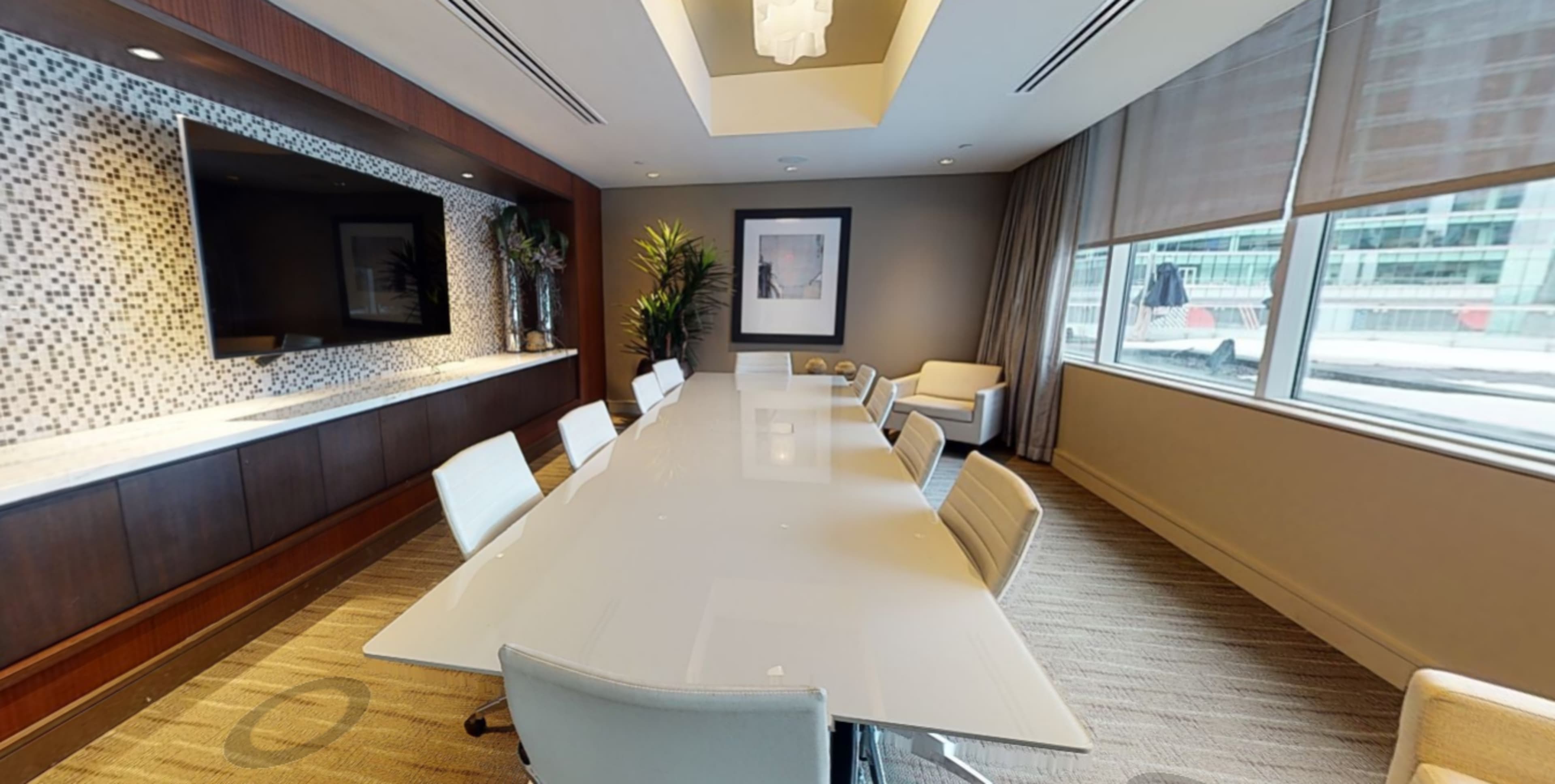 A modern conference room features a long white table surrounded by sleek chairs, with large windows providing natural light and a television mounted on the wall.