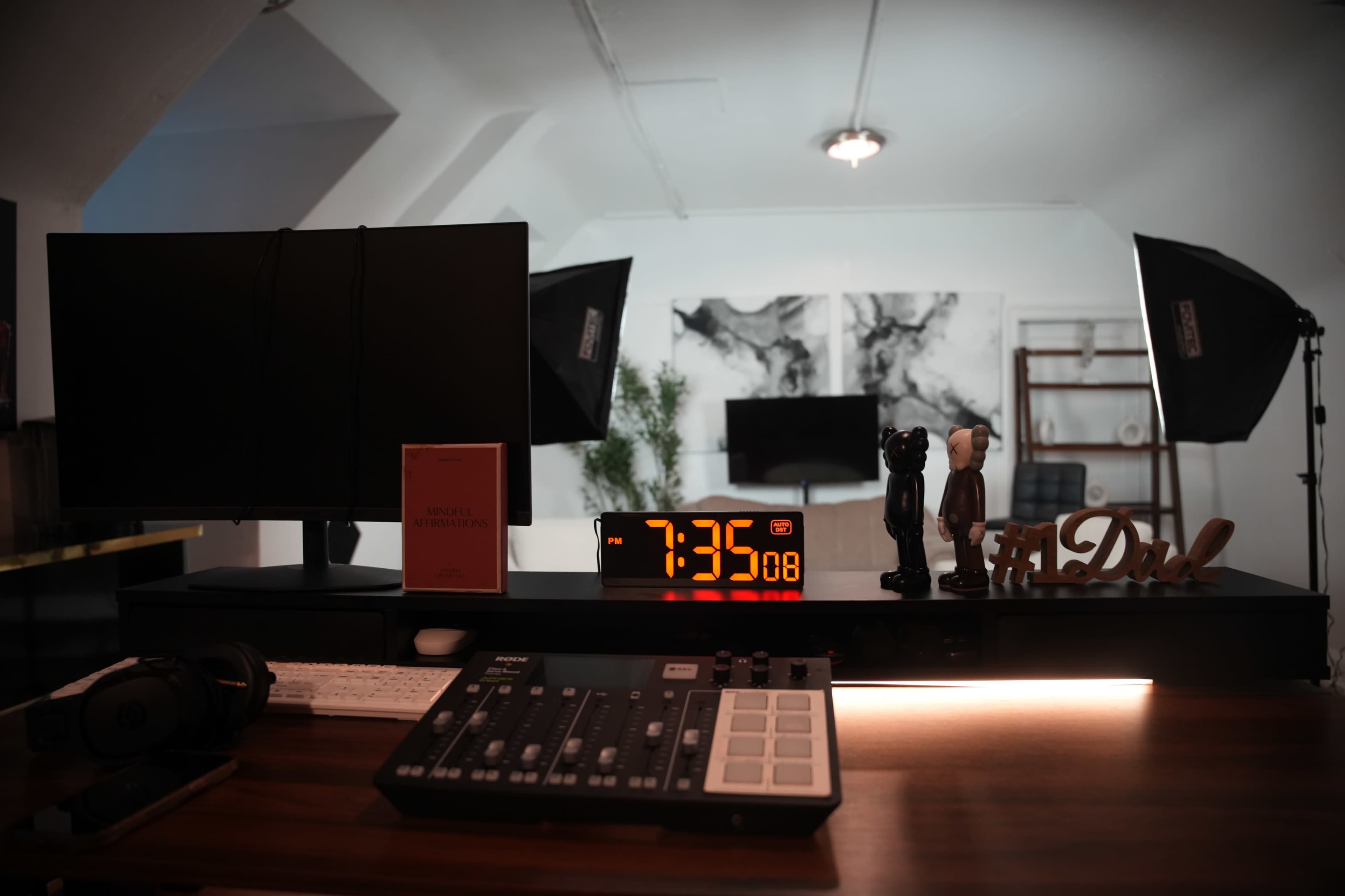 A modern workspace features two monitors, a digital clock displaying 7:35, a mixer, and decorative items on a desk.