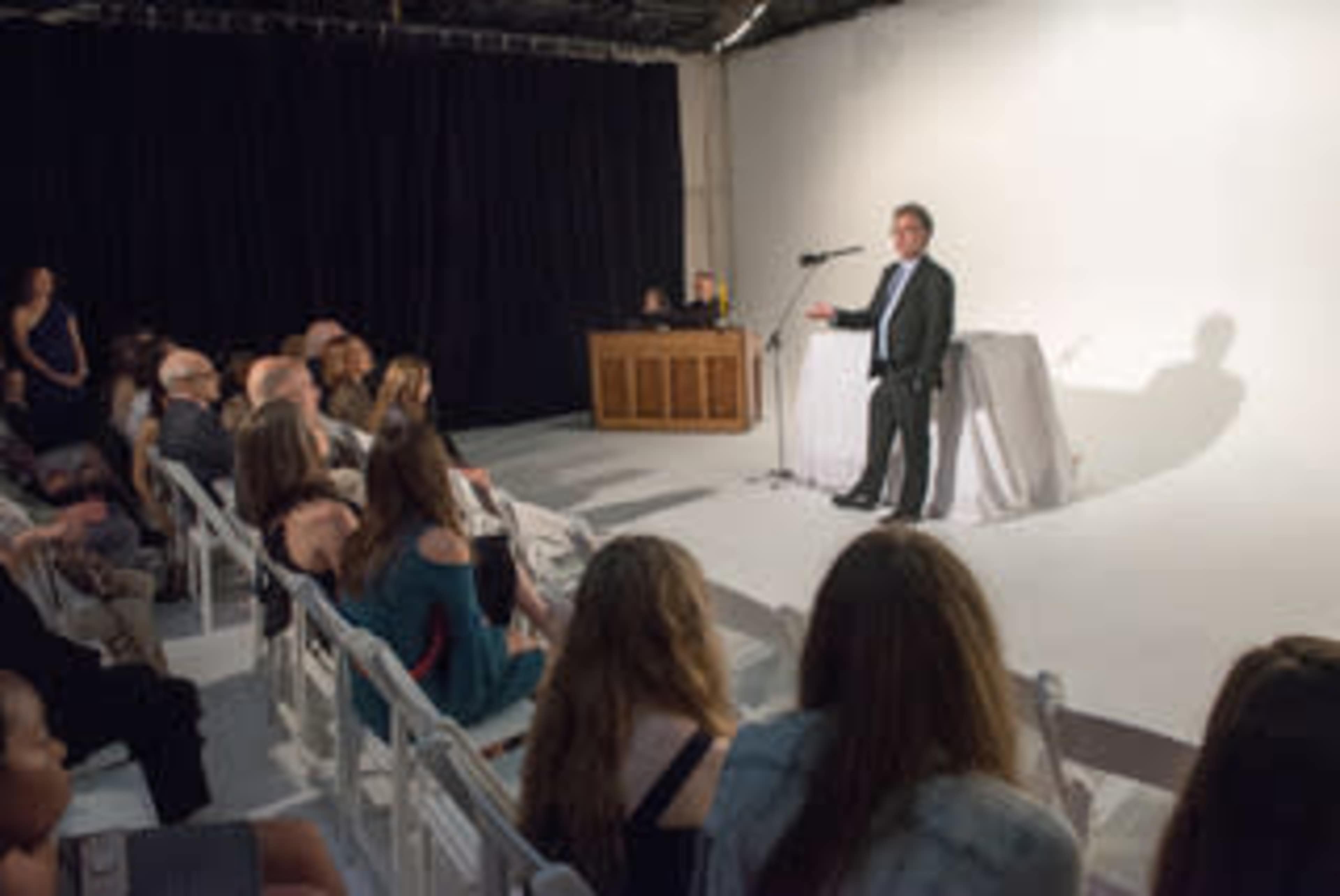 A speaker addresses an audience seated in rows, with a plain backdrop and a table in the foreground.