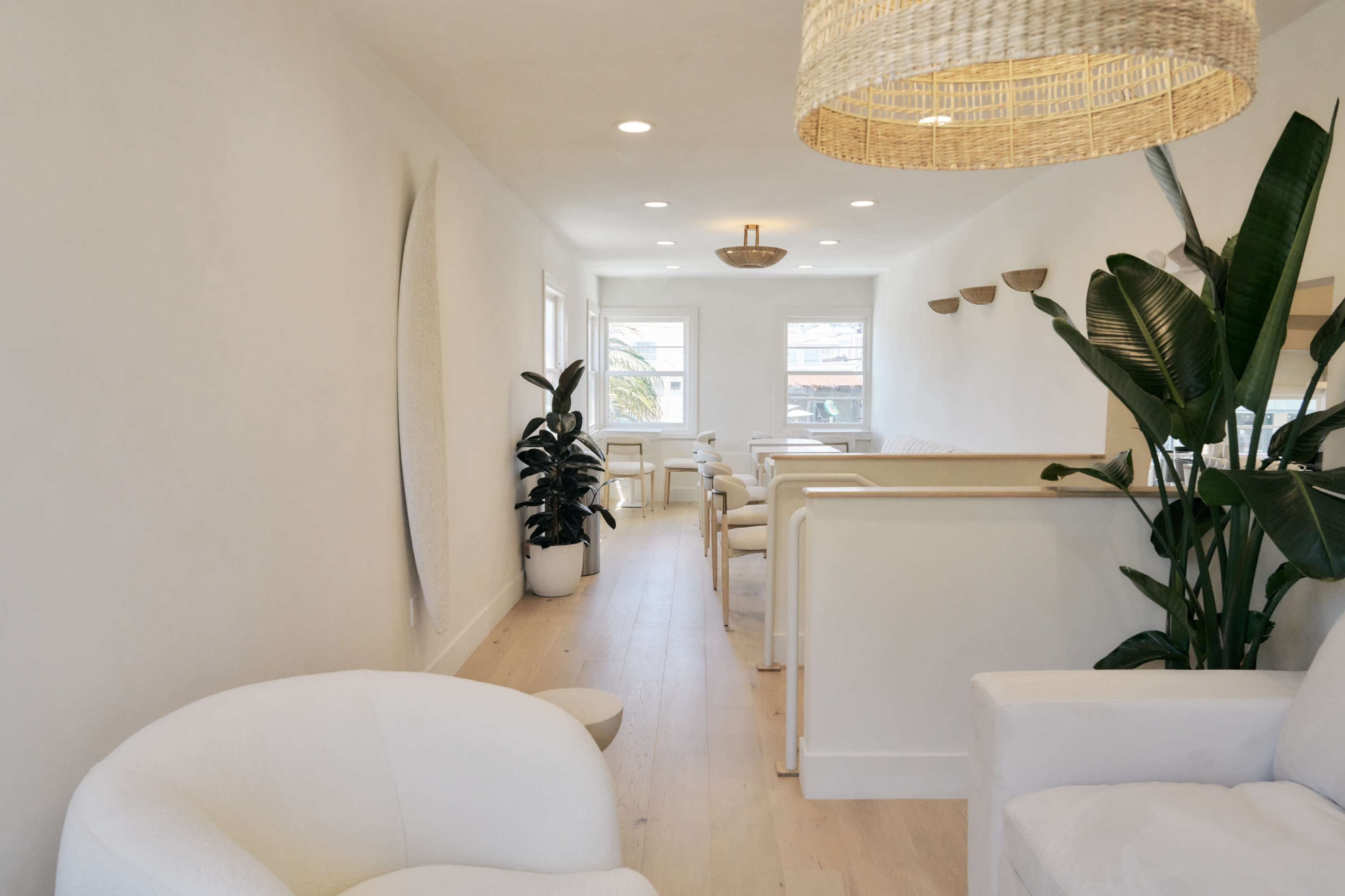 The image shows a bright, minimalist interior space with light wood floors, white walls, and modern furniture, featuring plants and a decorative light fixture.