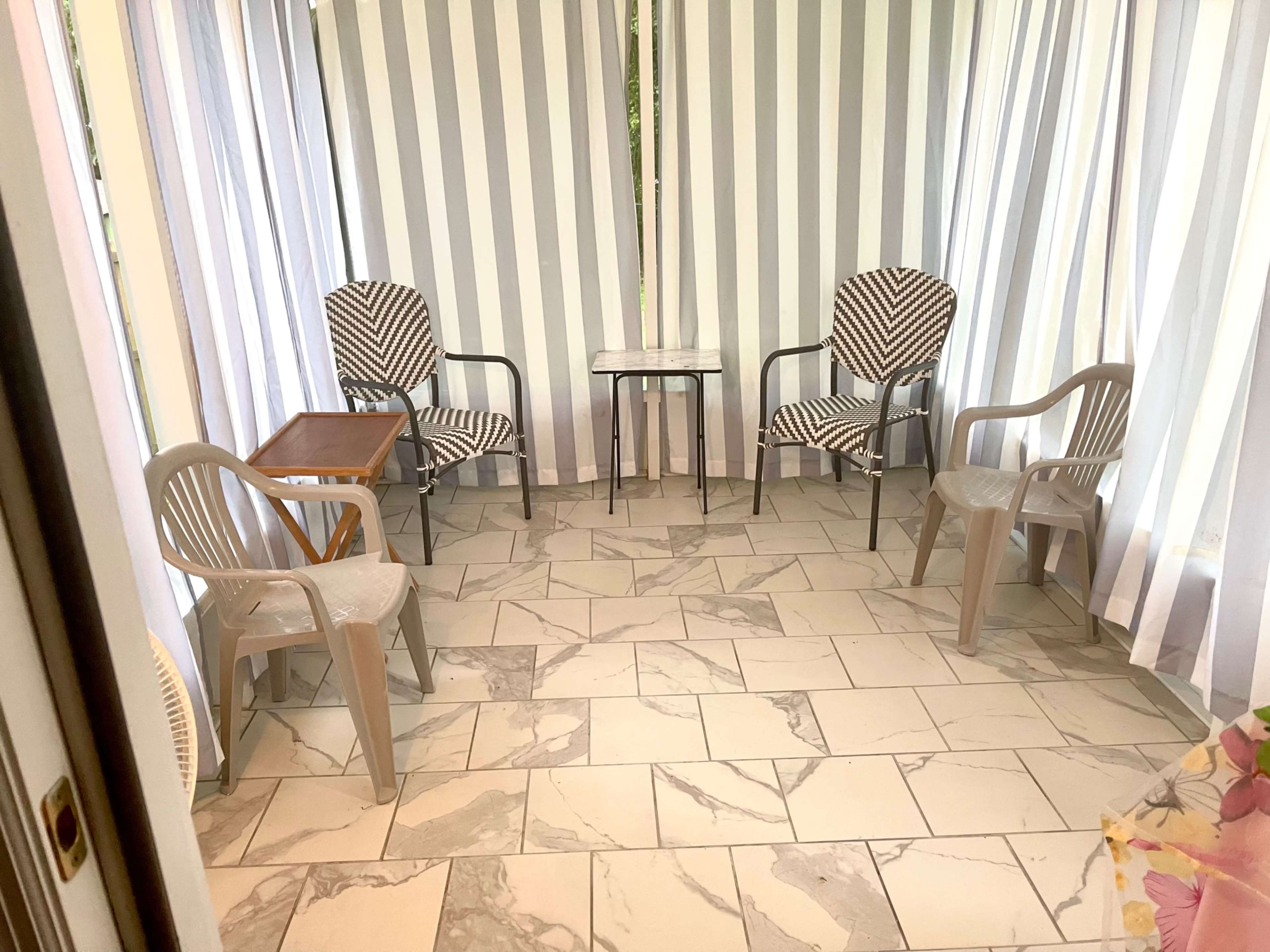 A sunroom with striped curtains, a marble-patterned floor, and various seating arrangements including two patterned chairs and a small table.