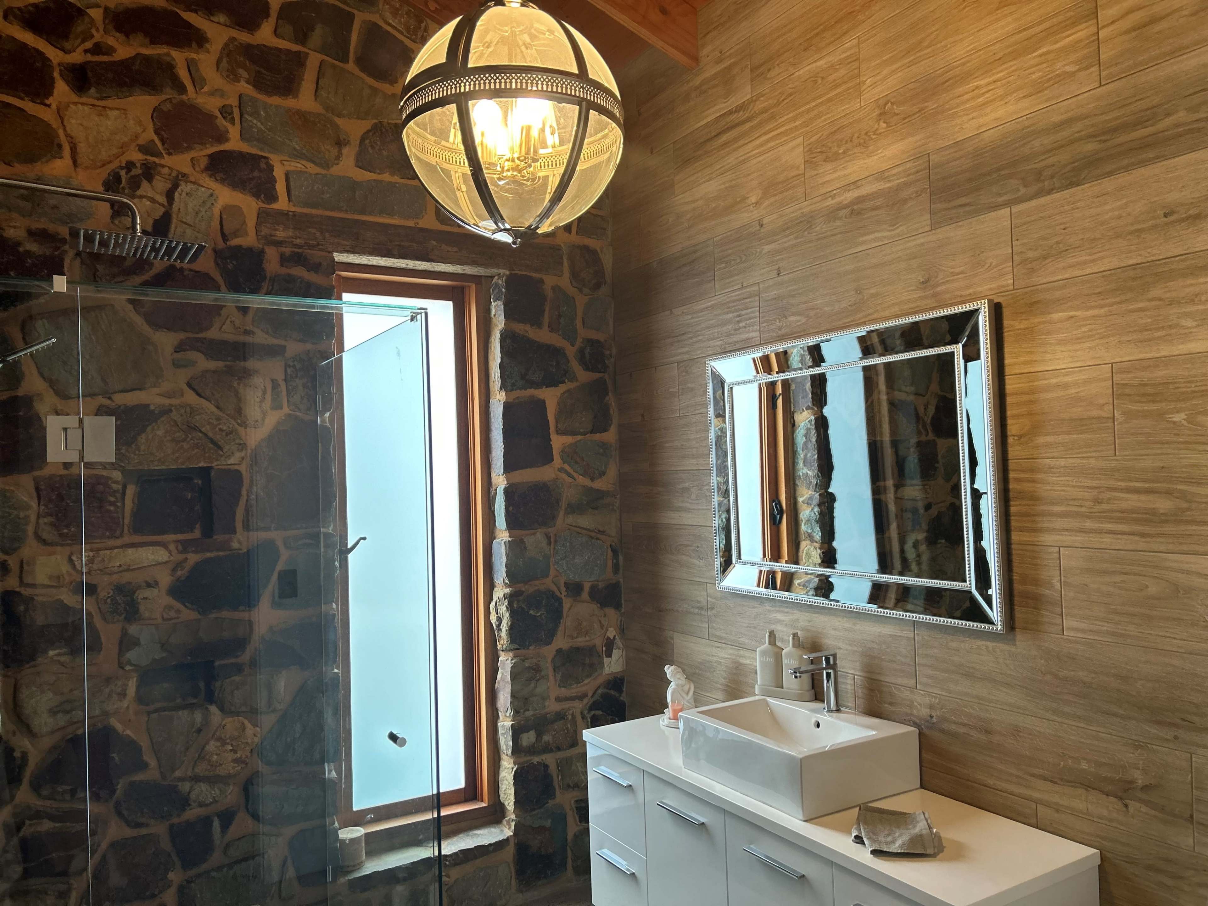 A modern bathroom featuring a glass shower, a stone wall, a wooden floor, a large mirror, and a decorative light fixture.