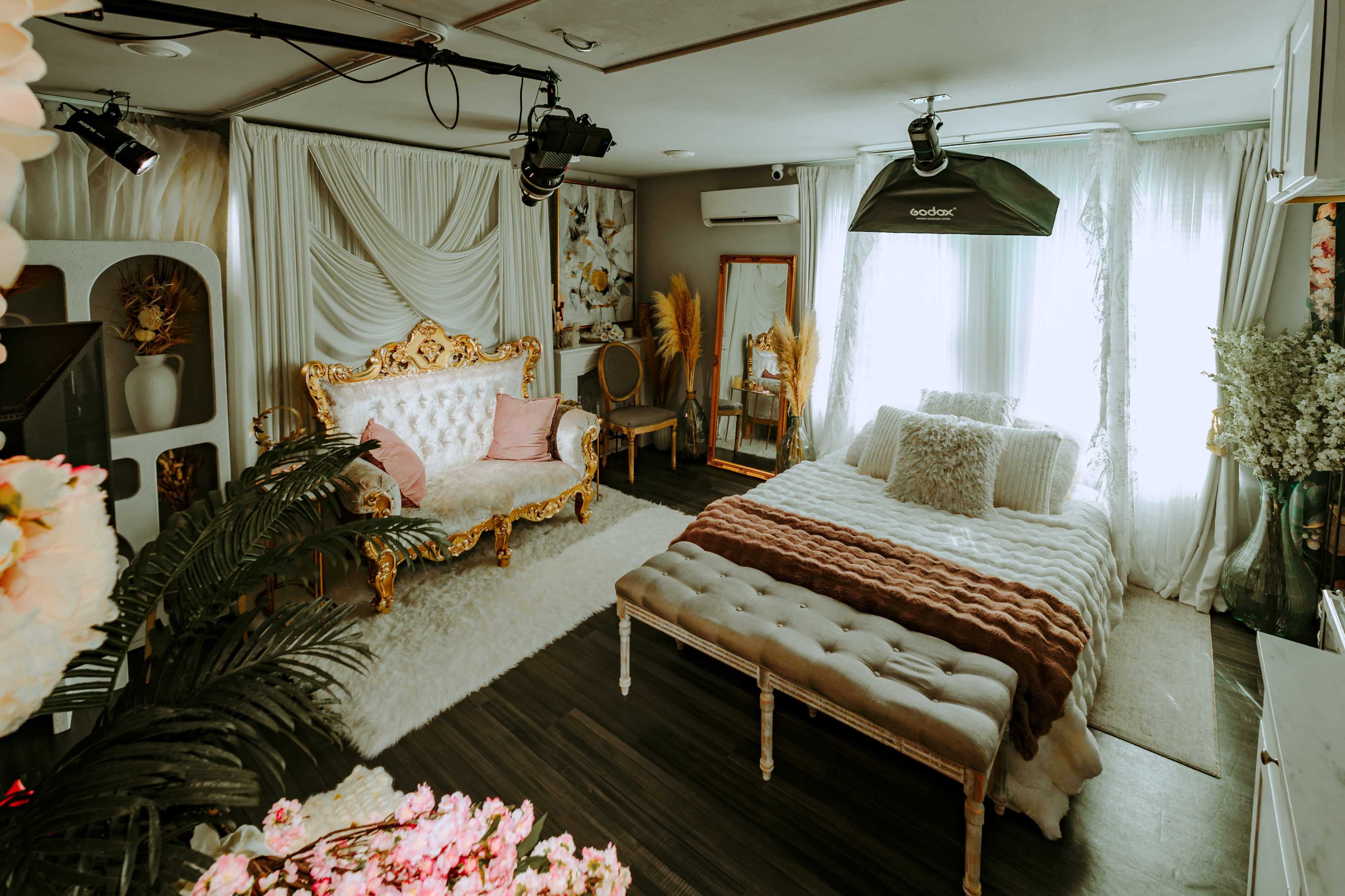 The image shows a decorated bedroom featuring a gold-accented sofa, a bed with a textured blanket, and soft, natural lighting from the windows.
