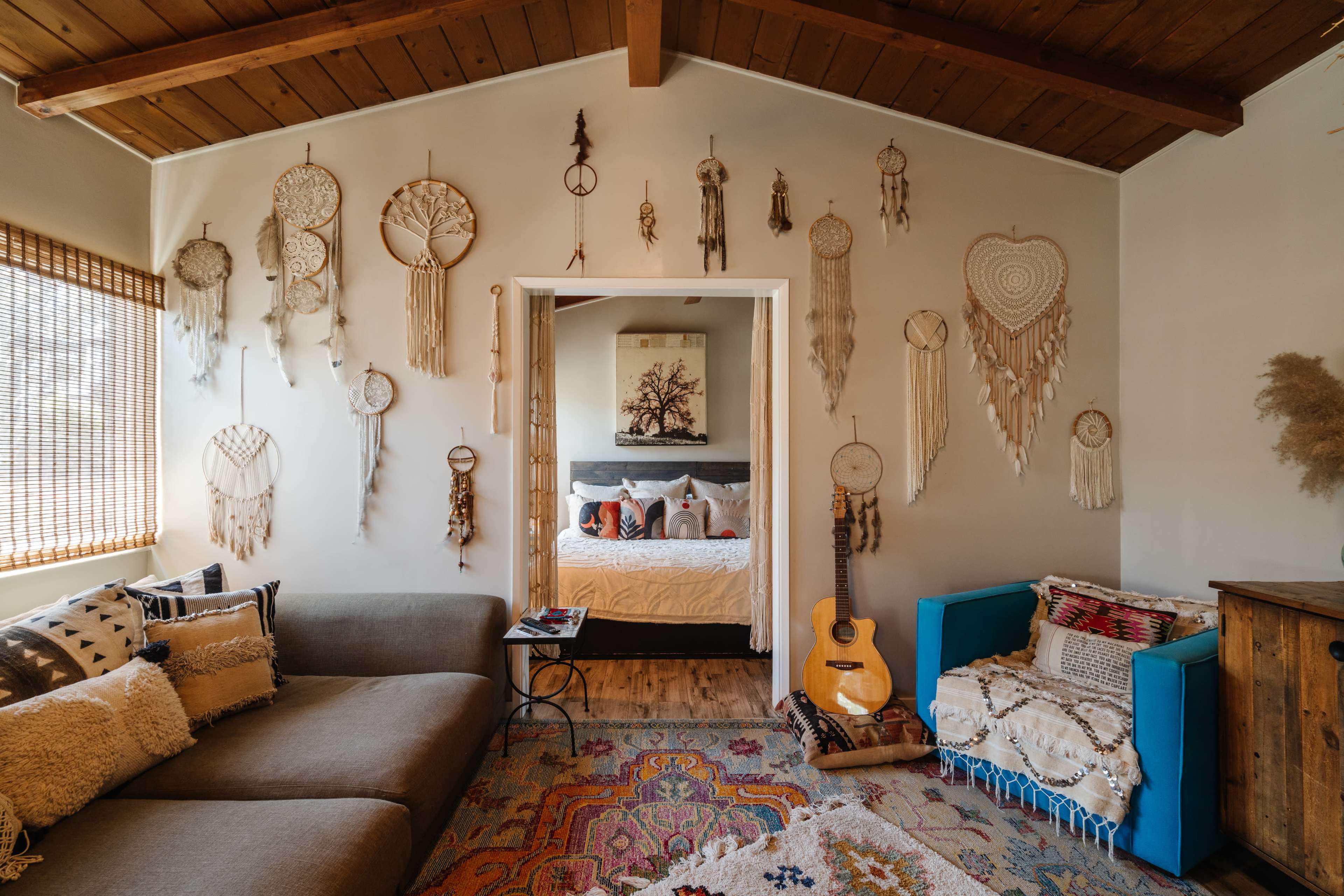 A cozy living room features a sectional sofa, colorful rugs, dreamcatchers on the walls, and an acoustic guitar resting beside a blue armchair.
