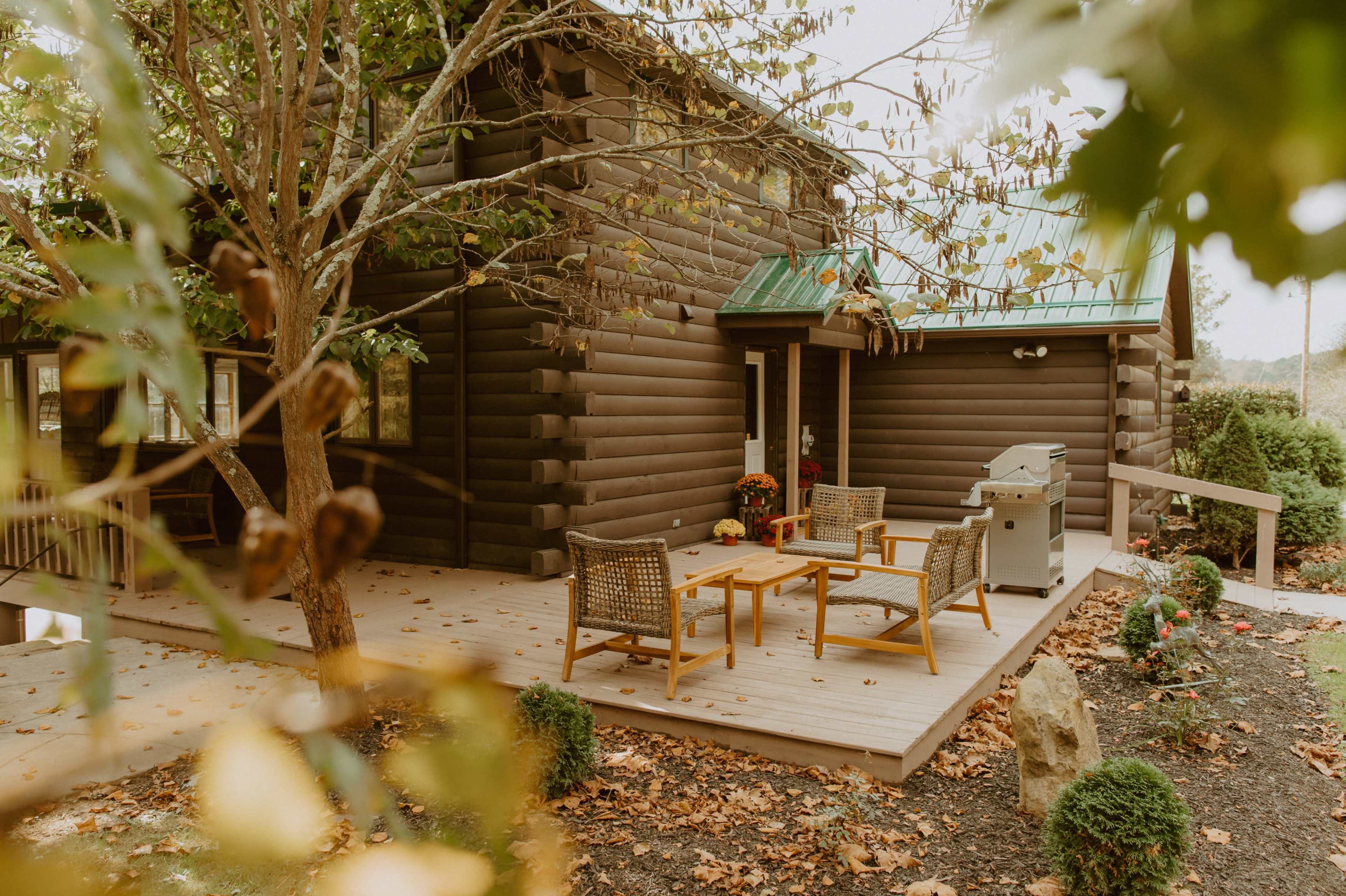 A wooden cabin features a patio with seating and a grill, surrounded by autumn leaves.