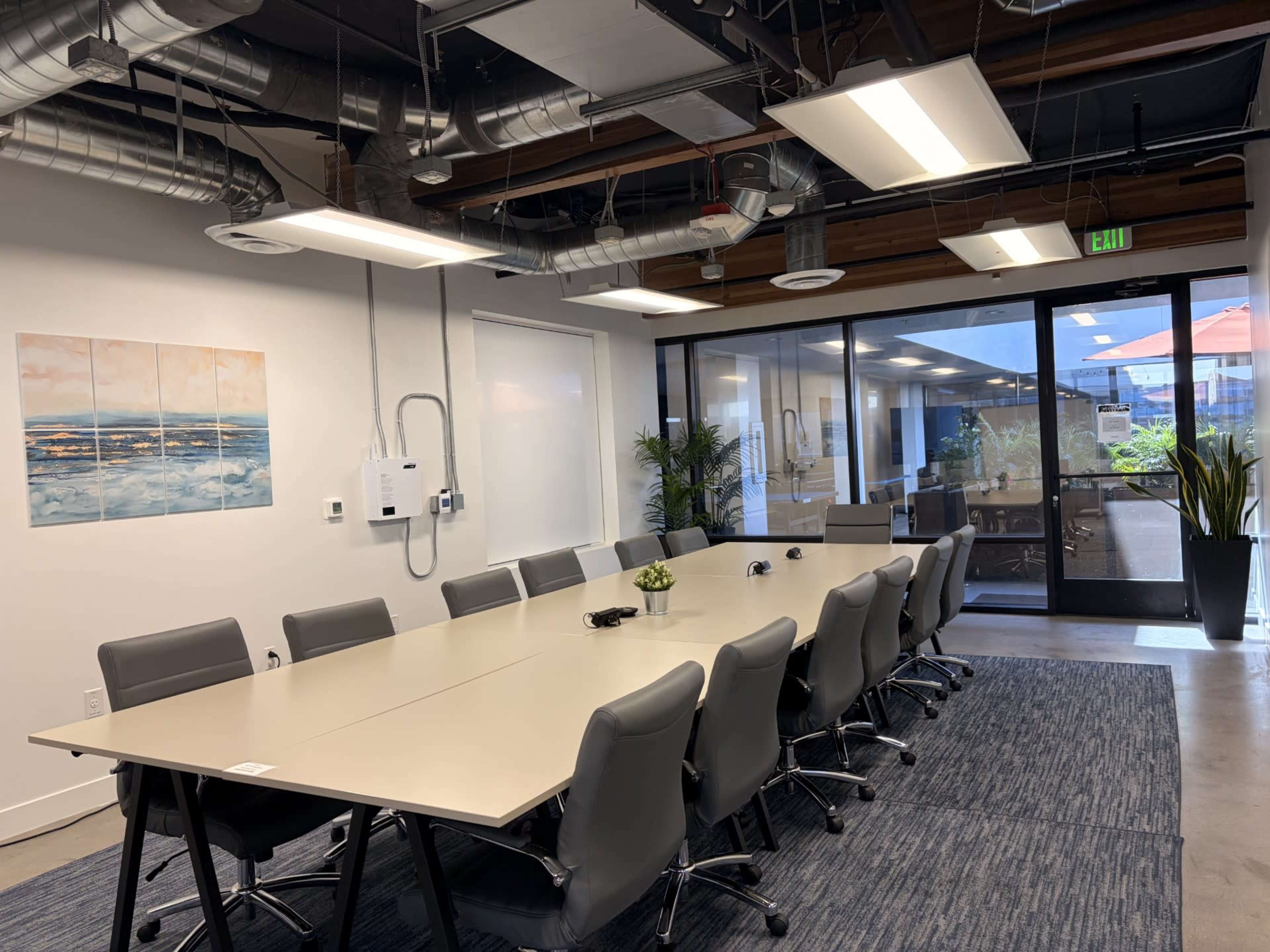 A modern conference room features a large table surrounded by gray chairs, with artwork on the wall and glass doors leading to an outdoor area.
