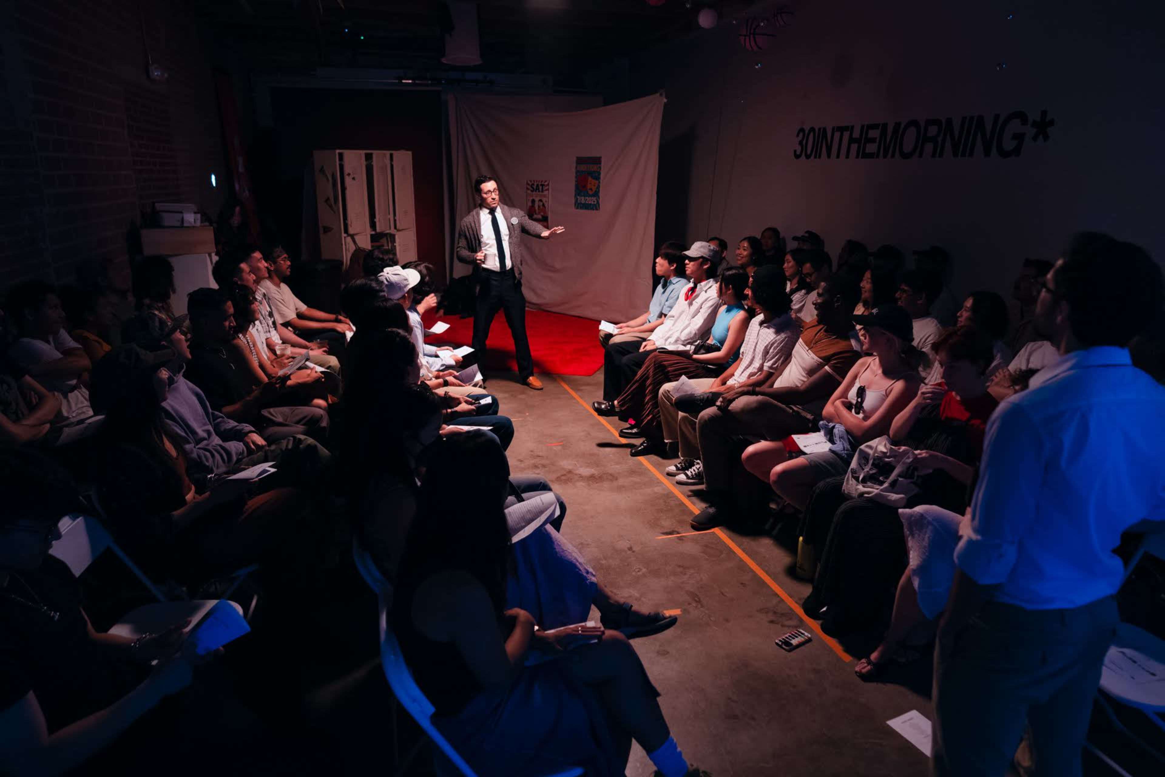 A speaker stands in front of a seated audience in a dimly lit room, with a backdrop featuring a white curtain and several pieces of art displayed on the walls.