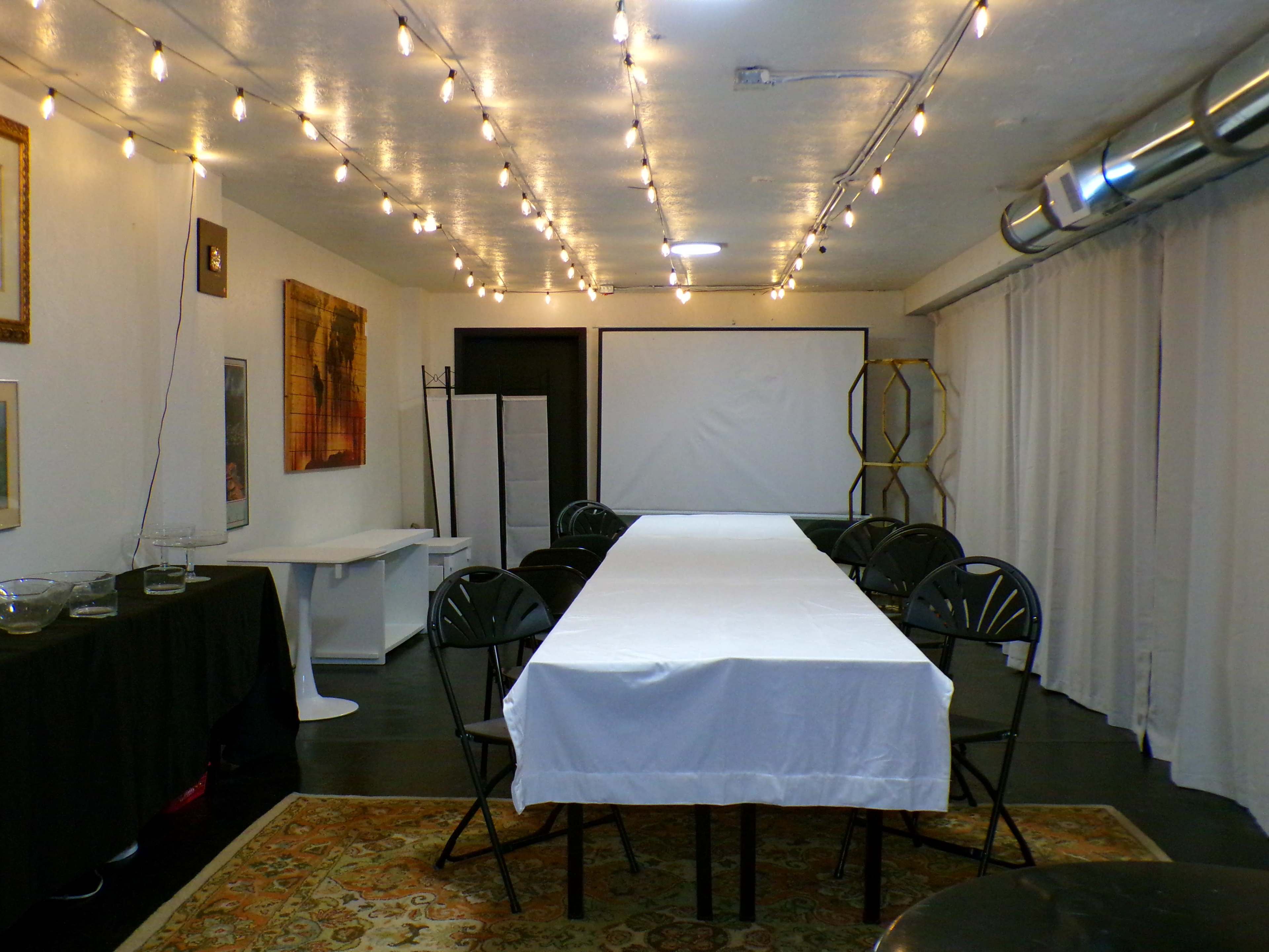 A spacious meeting room is set with a long white table surrounded by chairs, illuminated by string lights overhead, with a projector screen at one end.