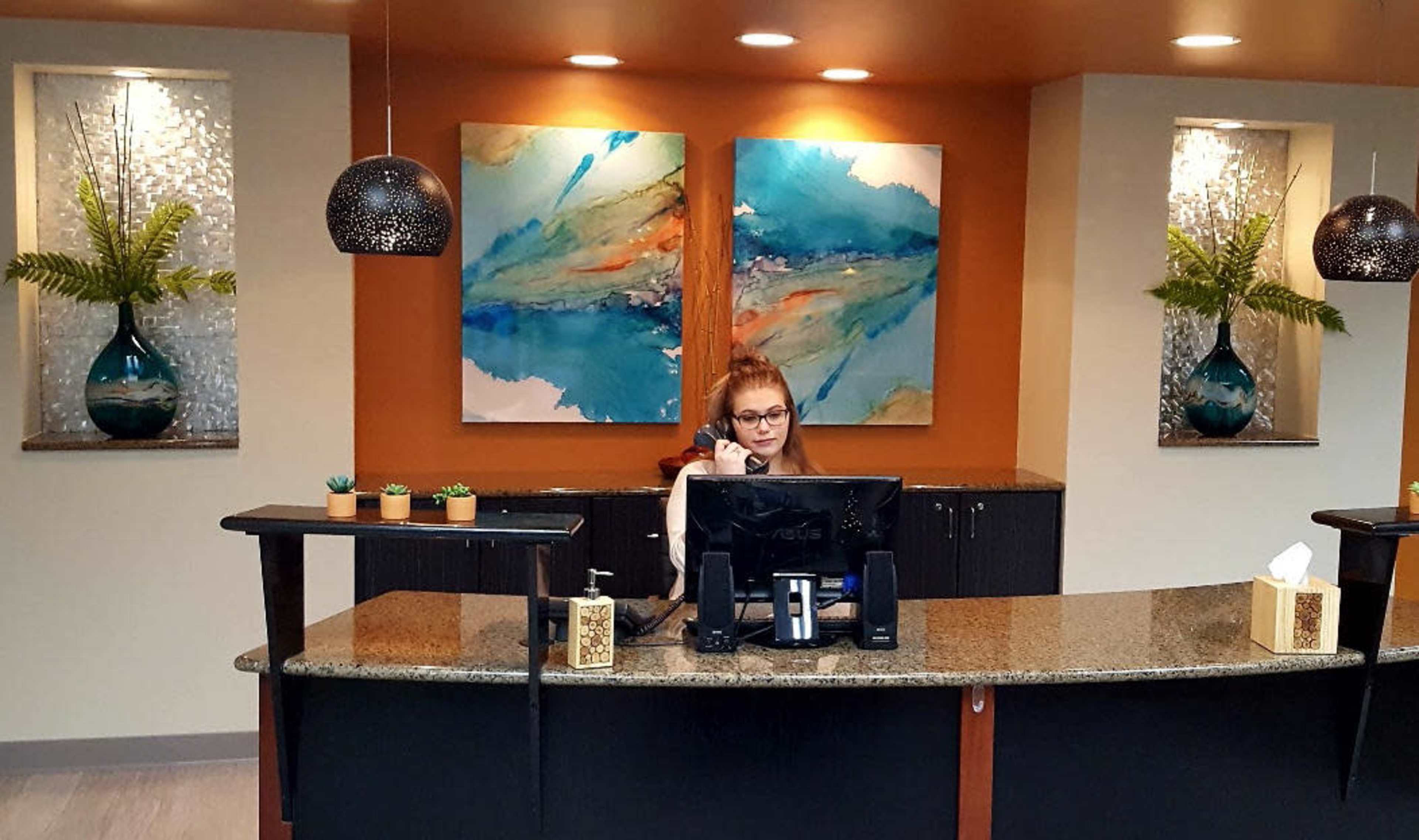 A woman sits at a reception desk, working on a computer, with abstract paintings and potted plants displayed on the wall behind her.