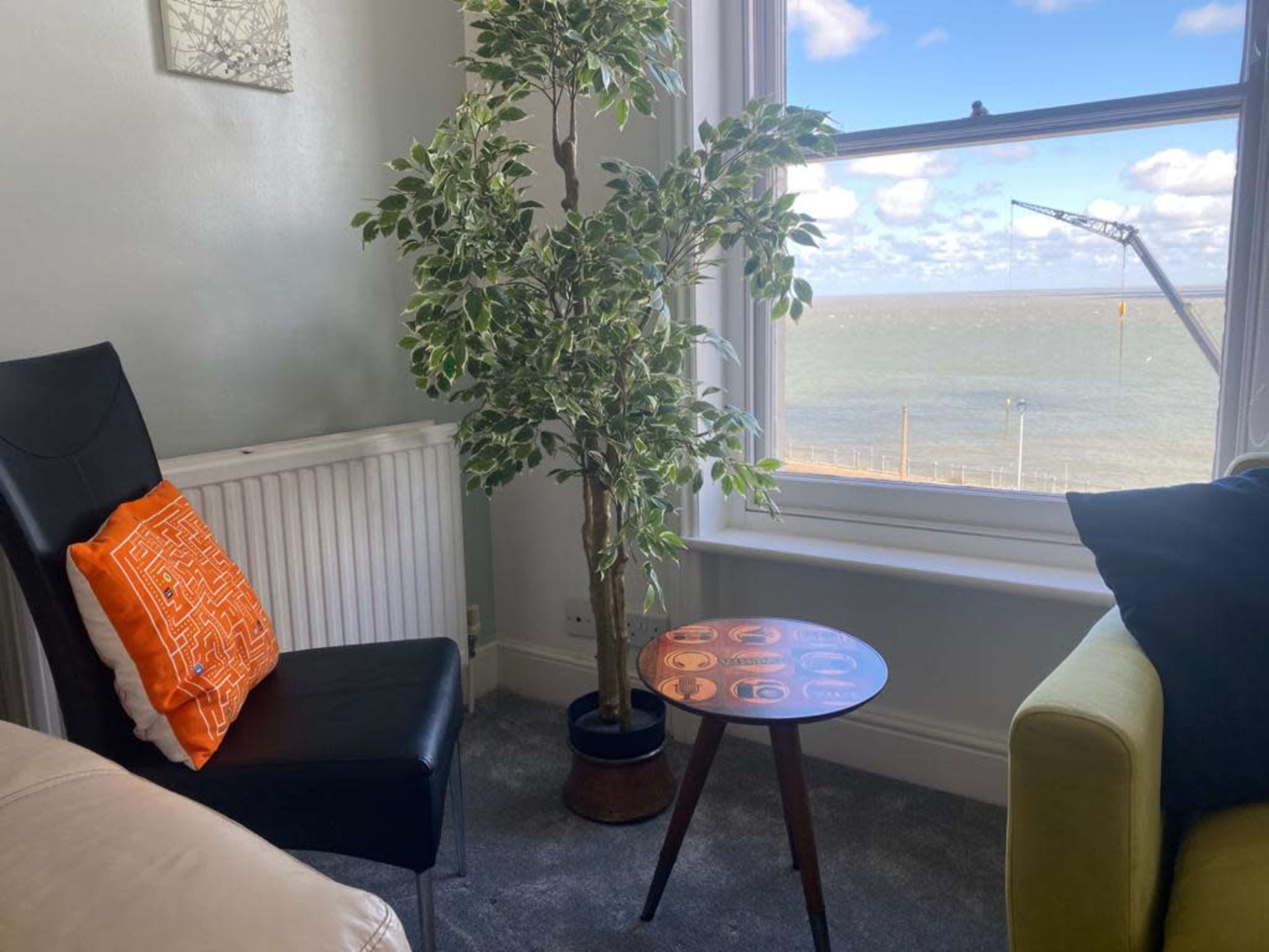 A corner of a room features a black chair with an orange pillow, a small round table, a potted tree, and a window showing a view of the sea and sky.