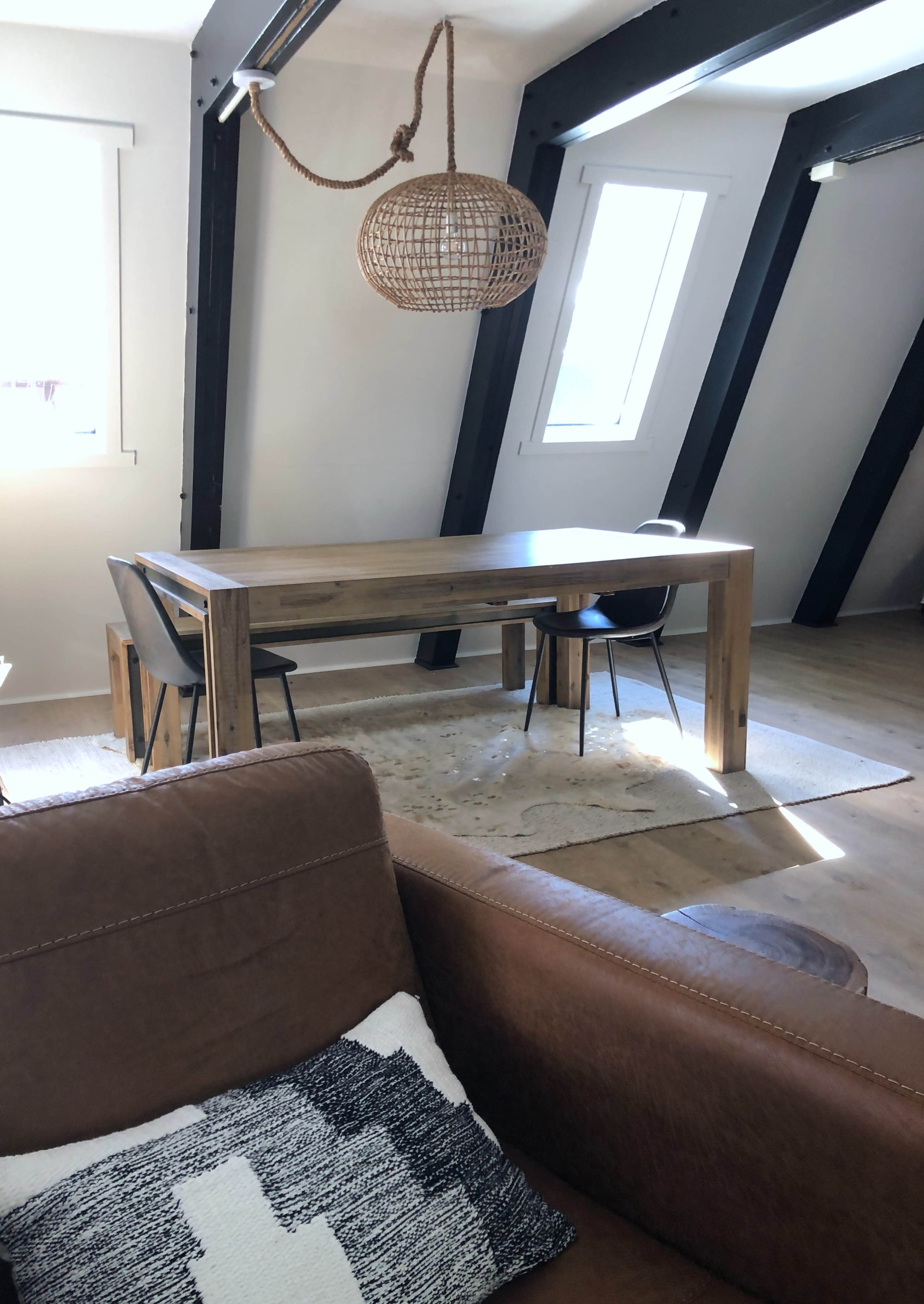A wooden dining table with two chairs is positioned in a well-lit room featuring exposed beams and a woven pendant light.