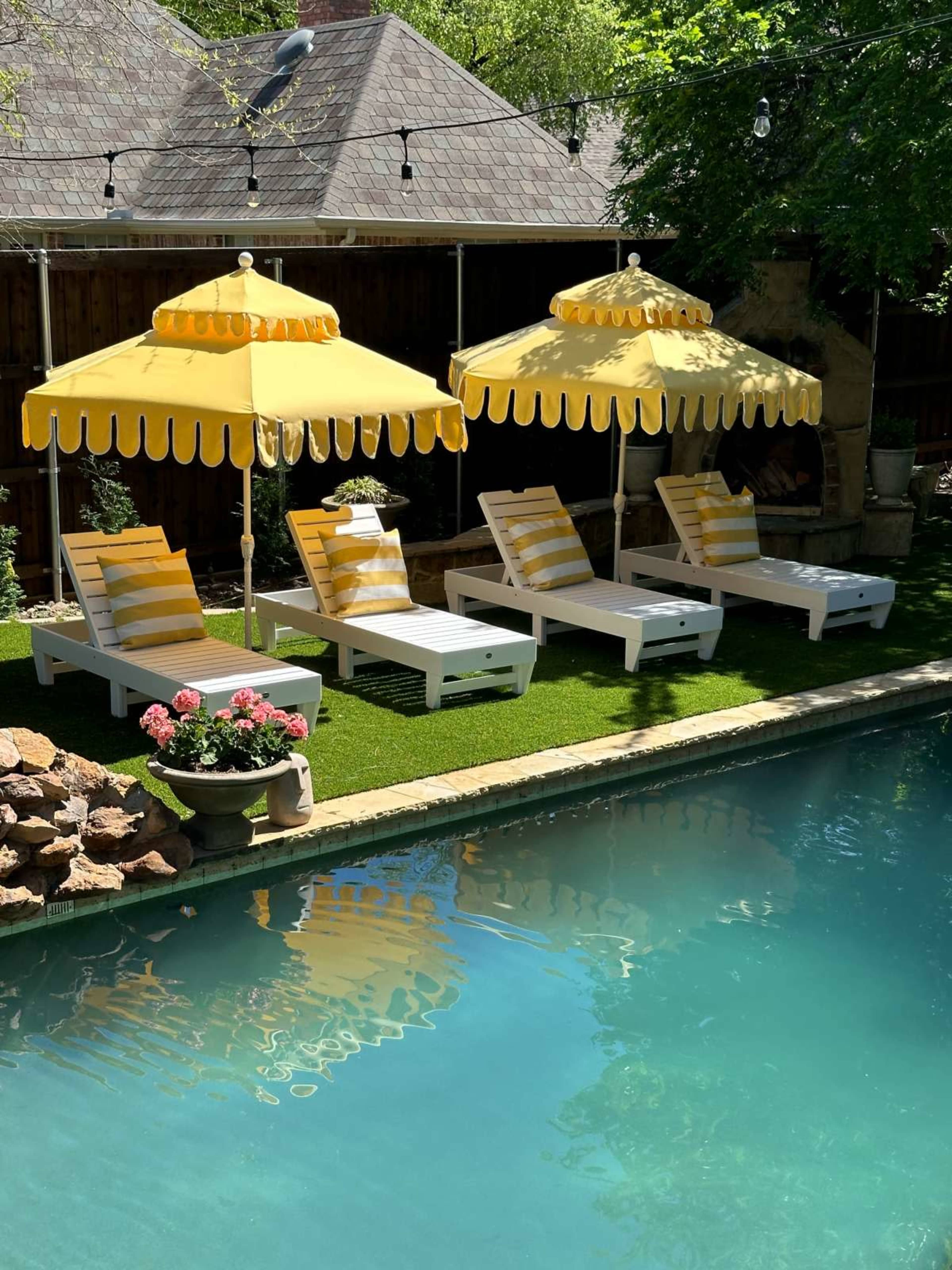 A backyard with four white lounge chairs under yellow-striped umbrellas by a swimming pool, surrounded by greenery and rocks.